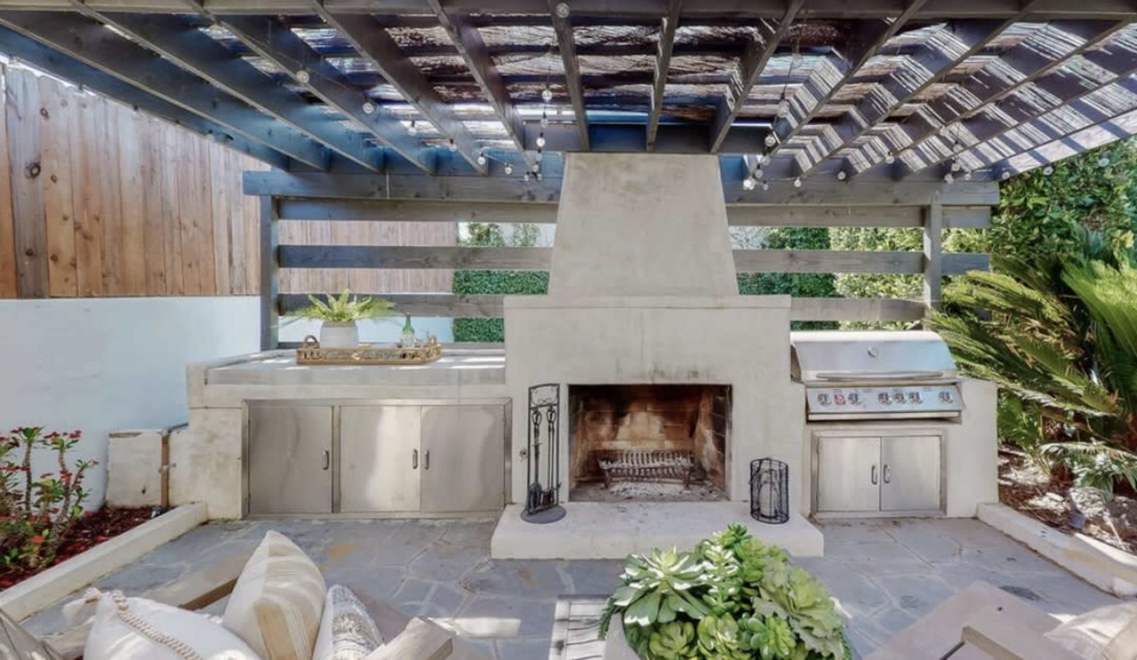The image shows an outdoor kitchen and dining area featuring a stone patio, a built-in grill, a fireplace, and a shaded pergola.