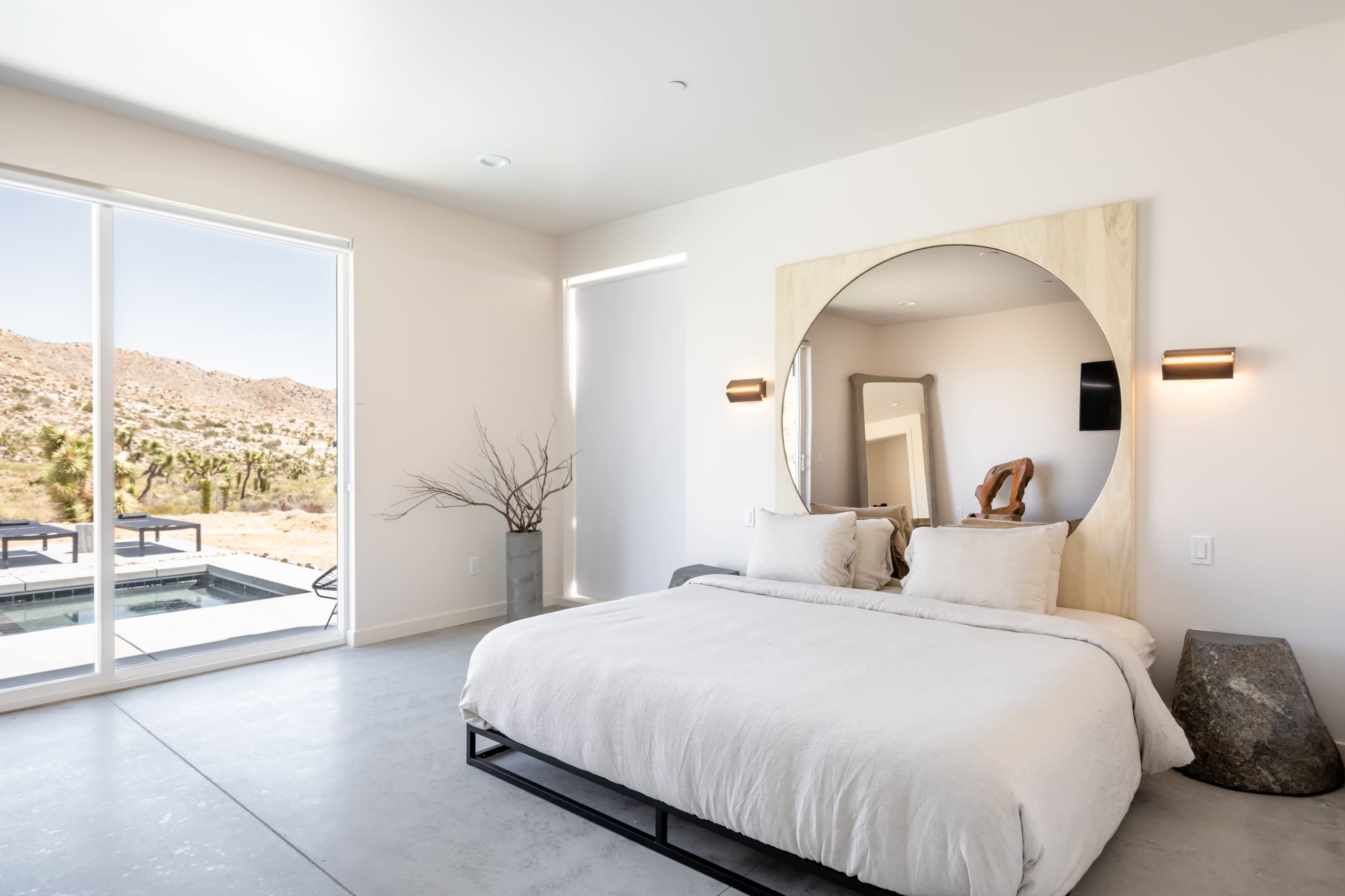 A minimalist bedroom features a large circular mirror above a bed with neutral bedding, large windows offering views of the desert landscape, and modern lighting fixtures.