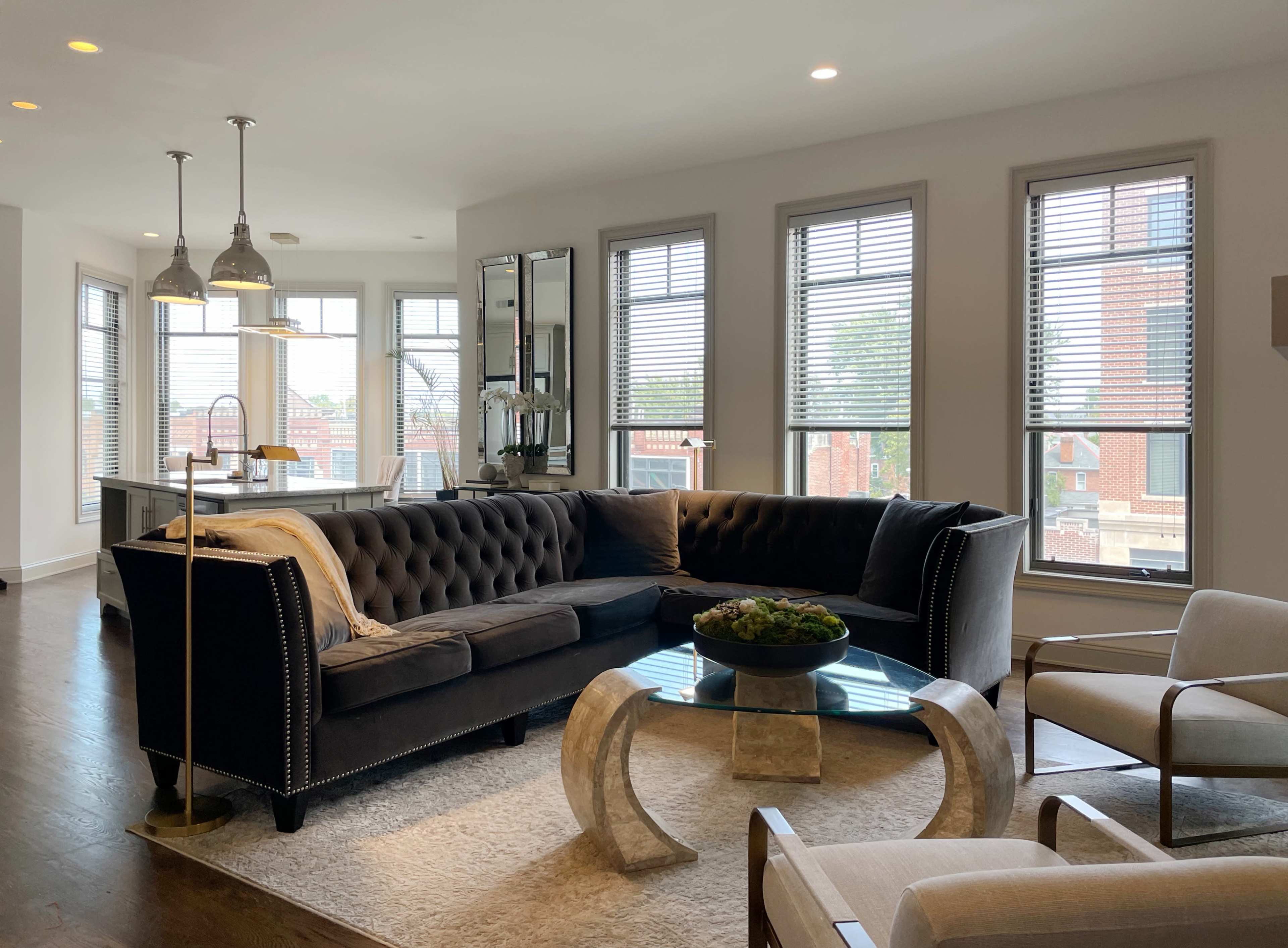 A modern living room with a large tufted sectional sofa, glass coffee table, and bright natural light streaming through multiple windows.