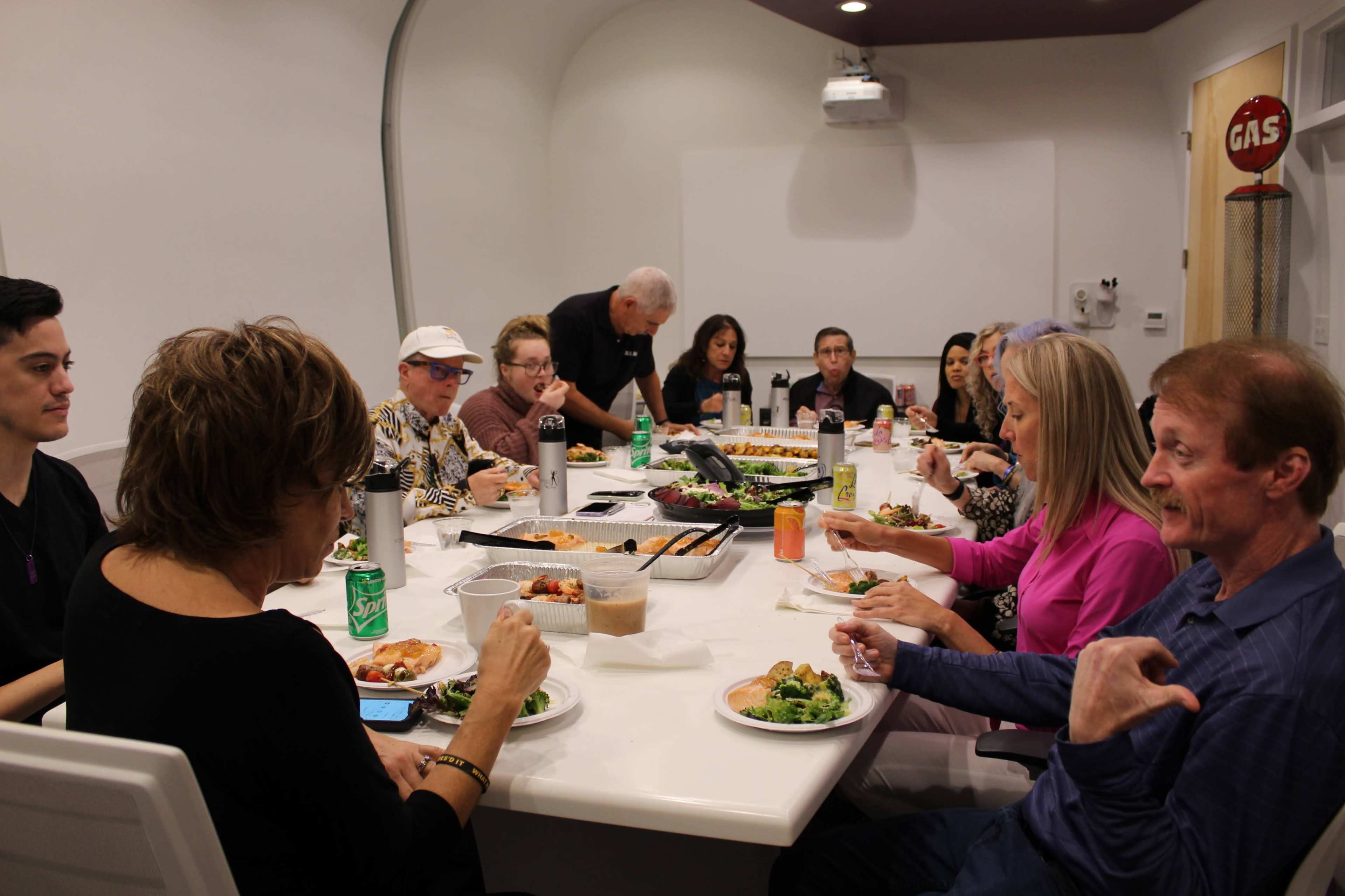 A group of people sits around a long table sharing a meal in a well-lit dining area.