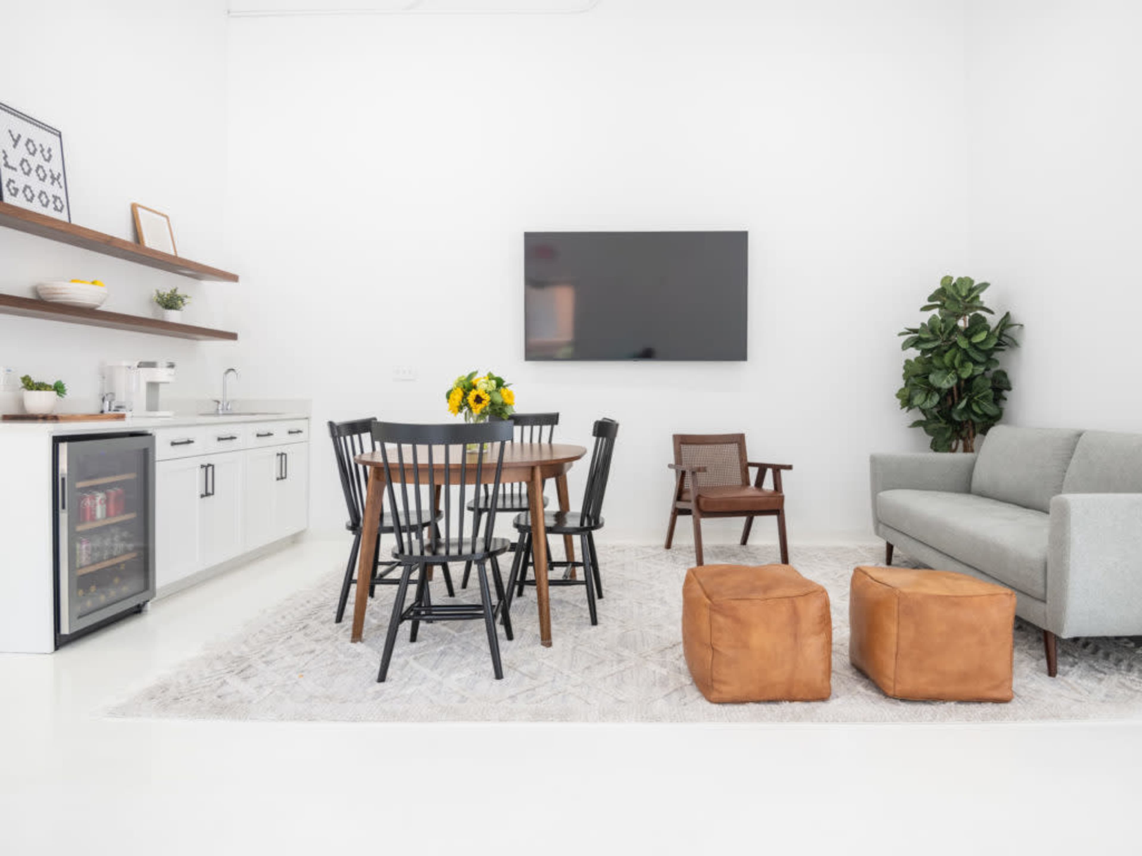 A modern, white-walled living space features a dining table with black chairs, a green sofa, a television on the wall, and a small kitchenette with a refrigerator.