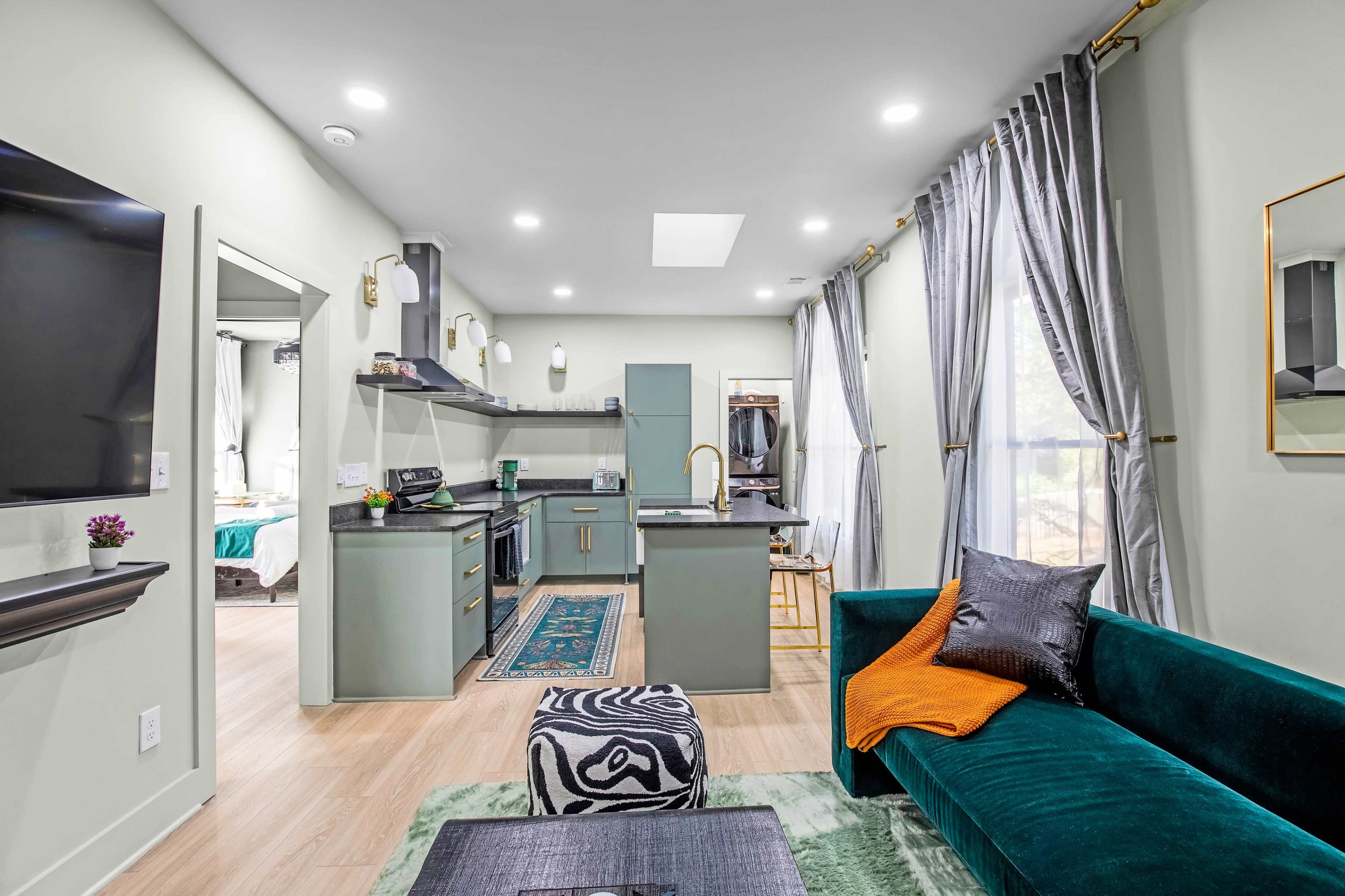 A modern living space featuring a green and gold kitchen, a dark green sofa, and large windows with sheer curtains.