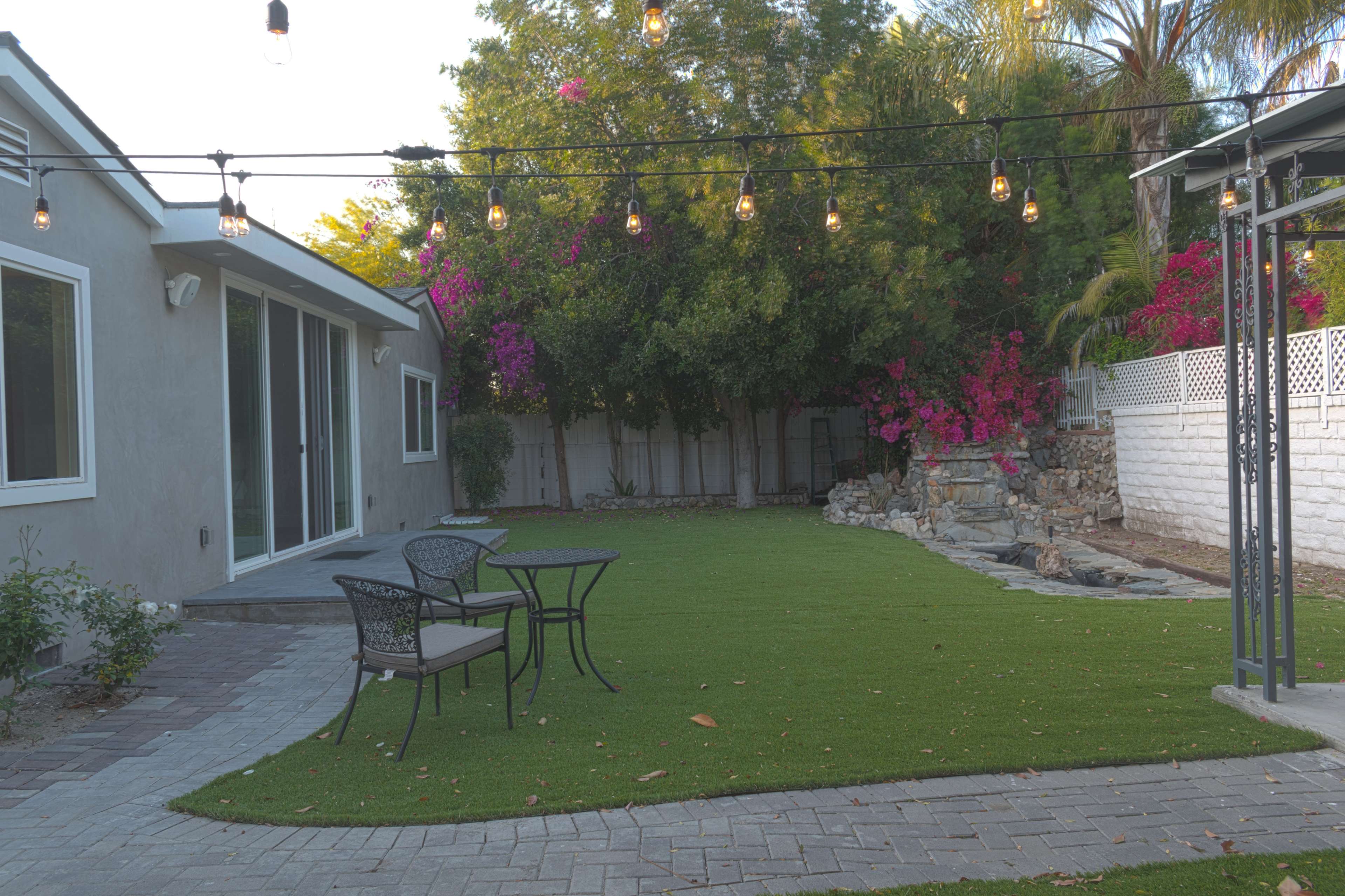 A patio area with a small table and chairs on artificial grass, surrounded by pink flowering bushes and a rock feature.