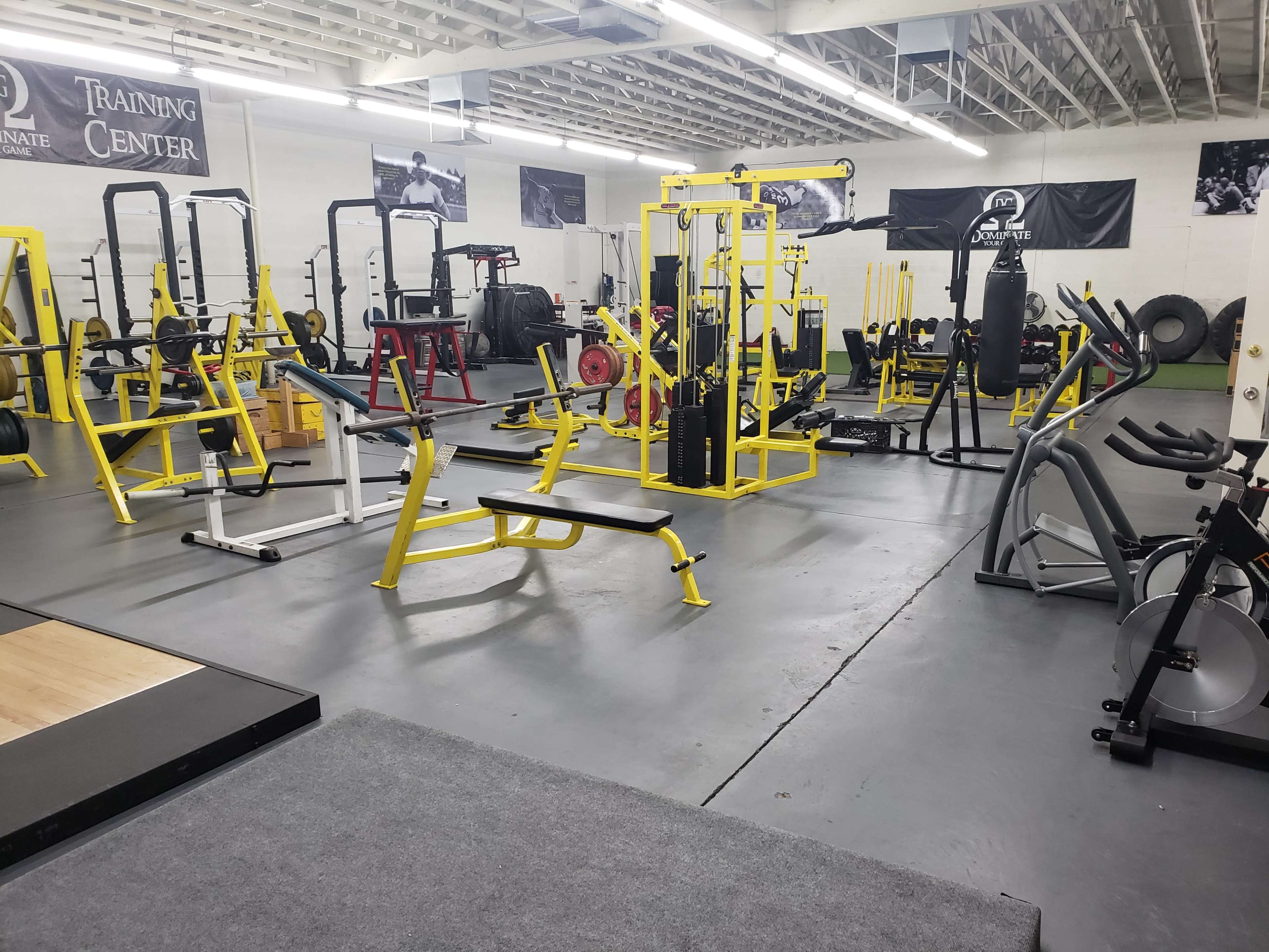 The image shows a spacious gym with various weightlifting and cardio equipment arranged on gray flooring.