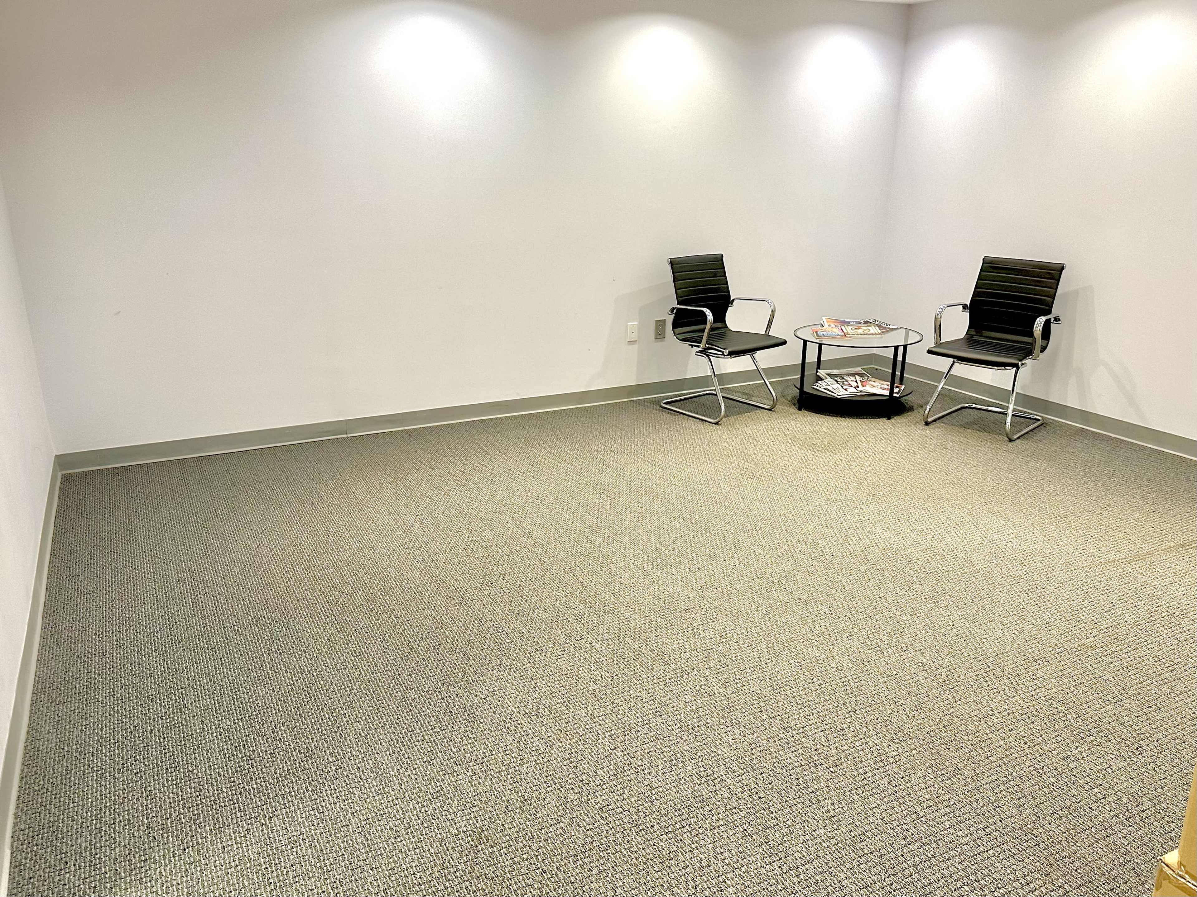 The image shows a small, empty room with two chairs and a round table in the center, set against a plain white wall.
