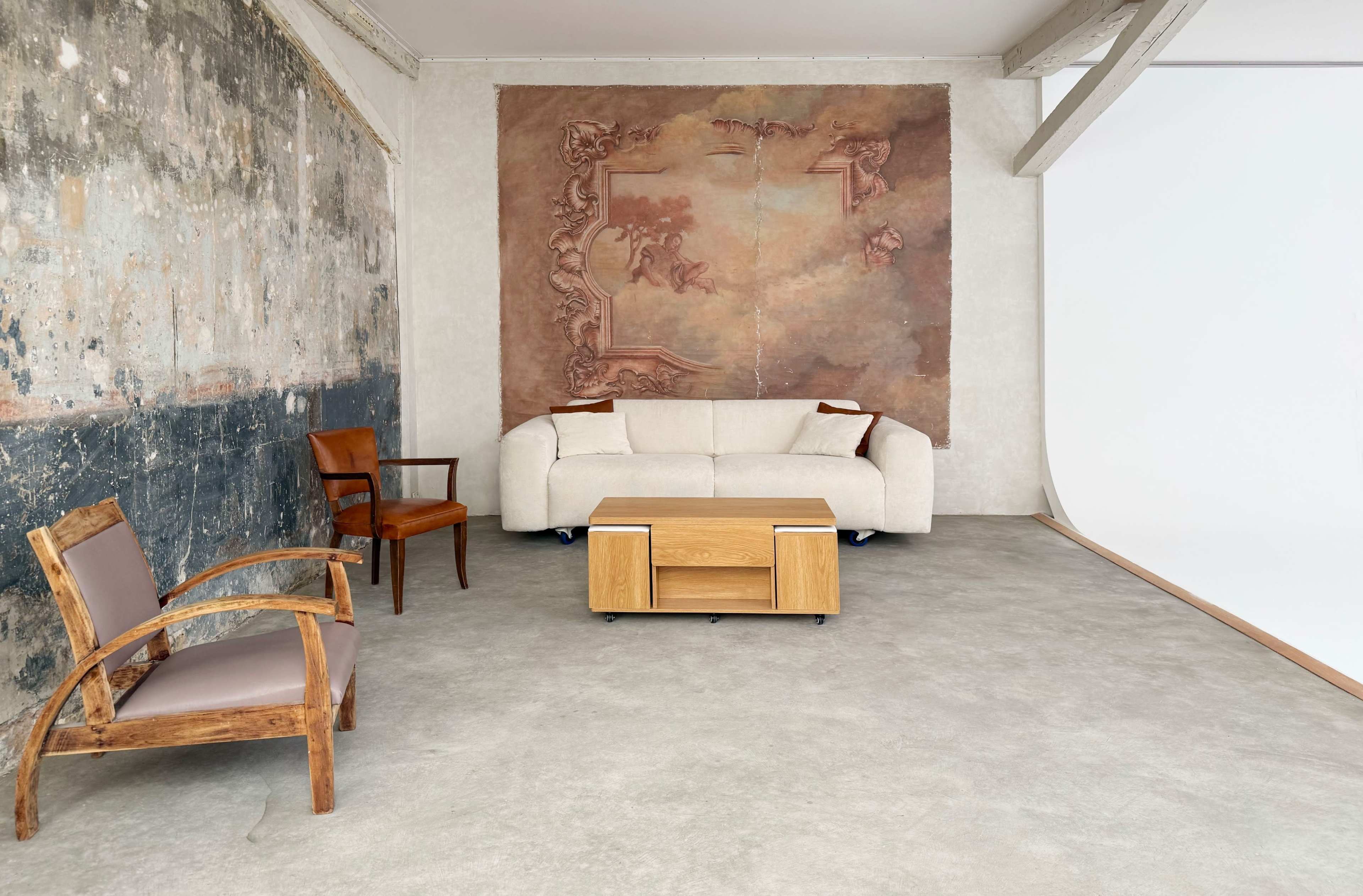 The scene features a minimalist interior with a light-colored sofa, a wooden coffee table, and a wooden chair, all set against a partially textured wall and an ornate mural.