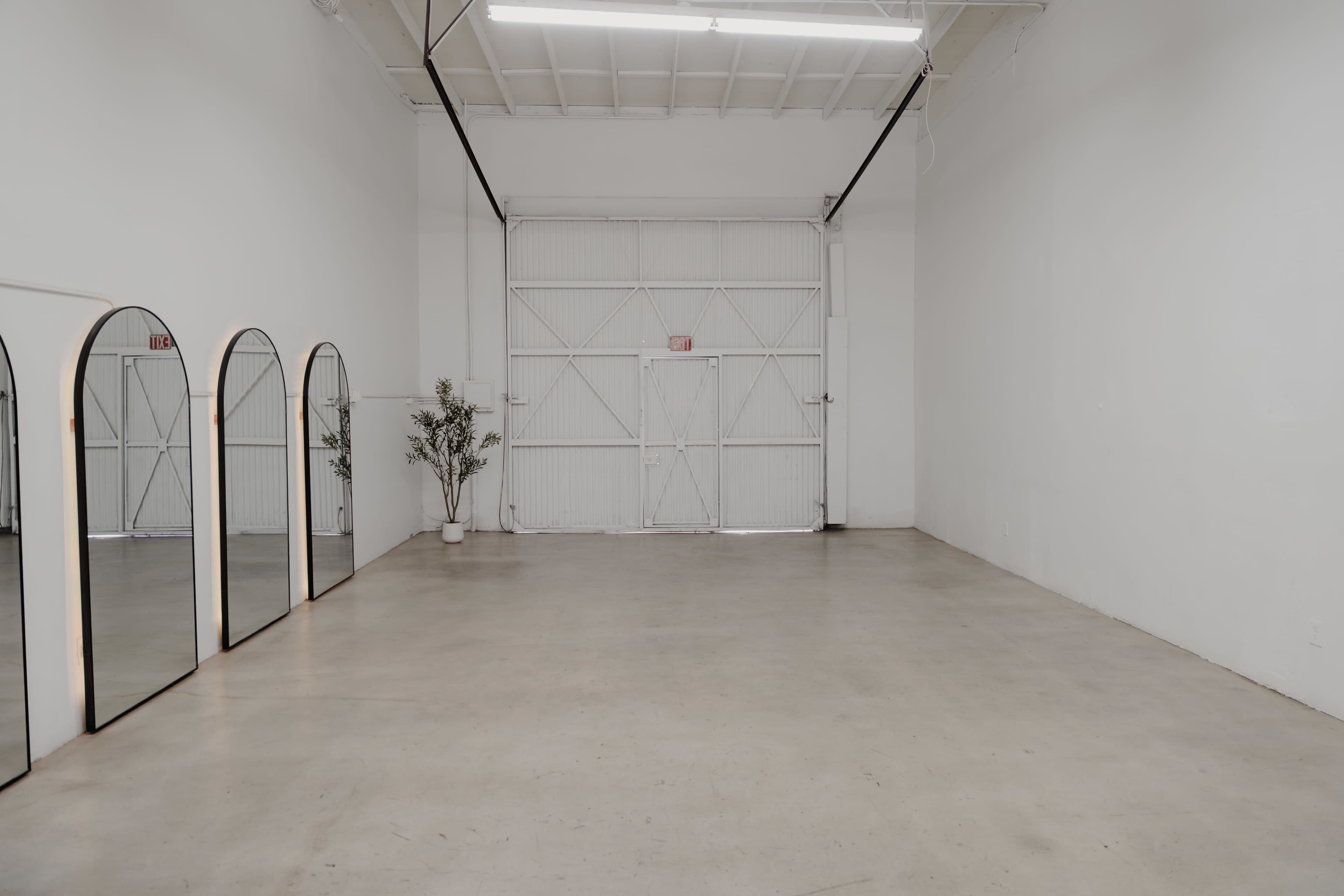 The image shows an empty room with white walls, a concrete floor, a large set of double doors at one end, and four arched mirrors on one side.