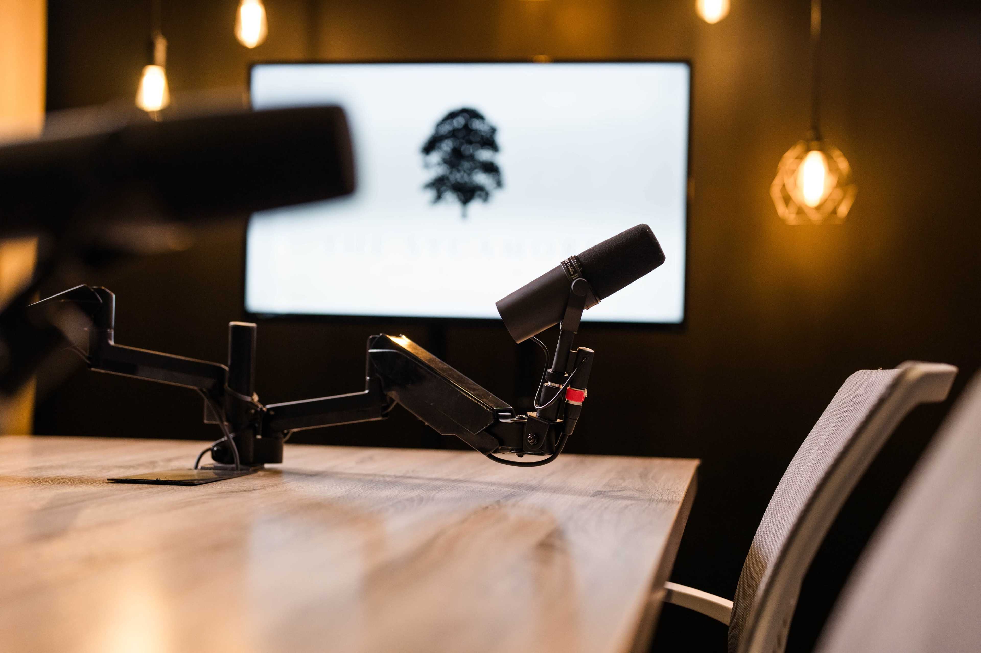A tabletop setup features microphones on adjustable arms, with a television screen displaying a logo in the background.