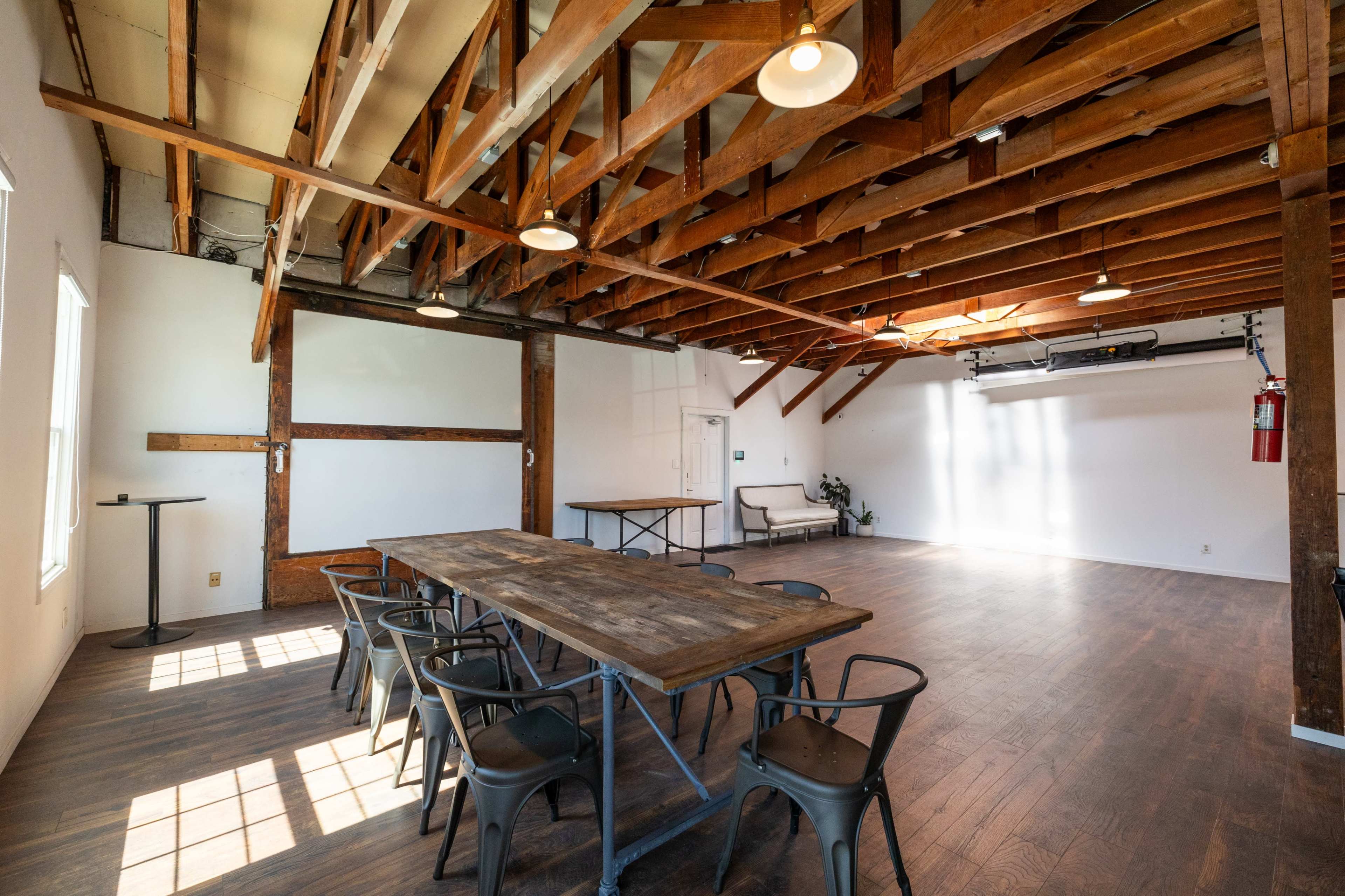 The scene shows a spacious, well-lit room with wooden beams, a large table, several chairs, and a fire extinguisher on the wall.
