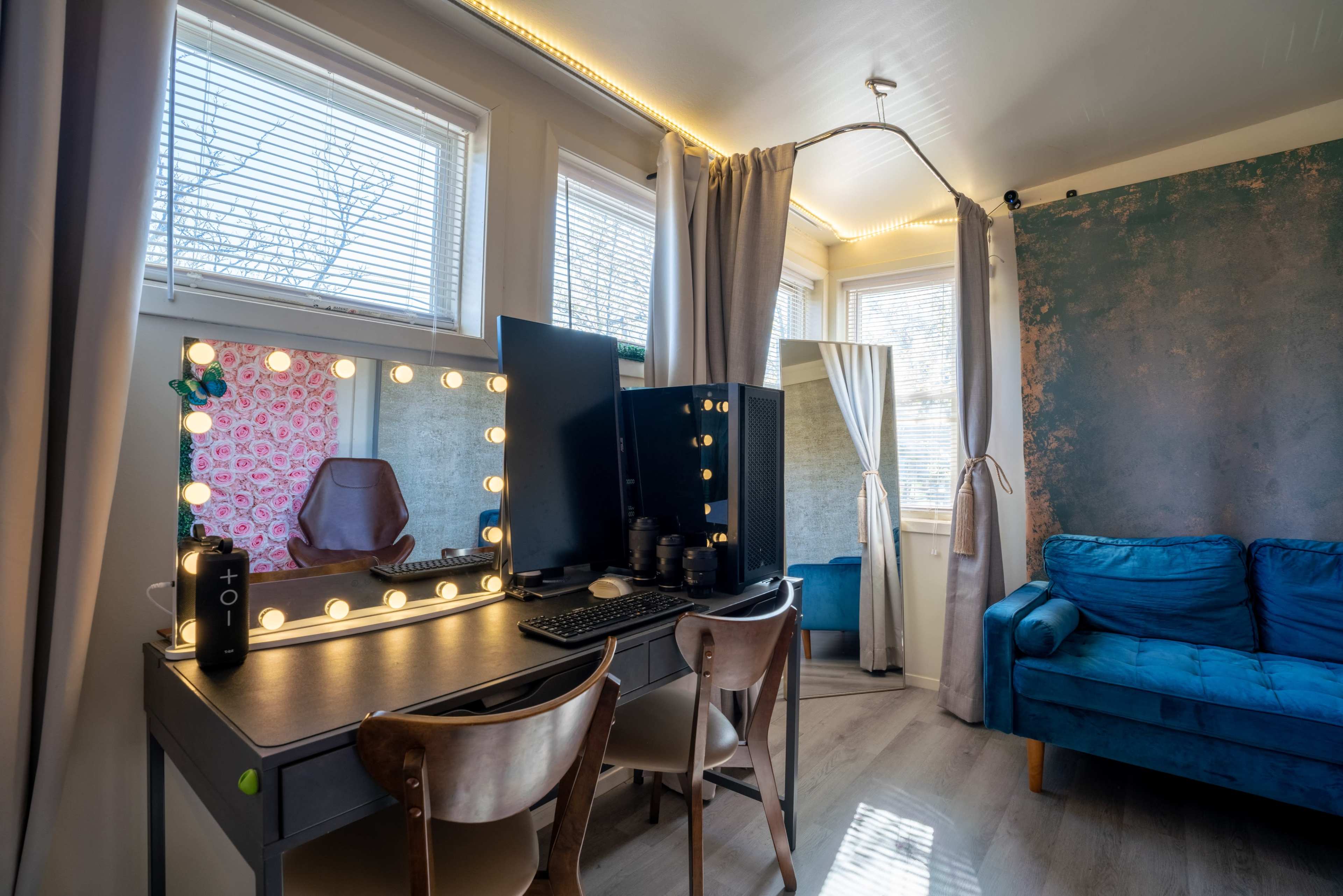 A brightly lit room featuring a desk with a mirror adorned with lights, a computer monitor, and a blue couch next to a window with curtains.