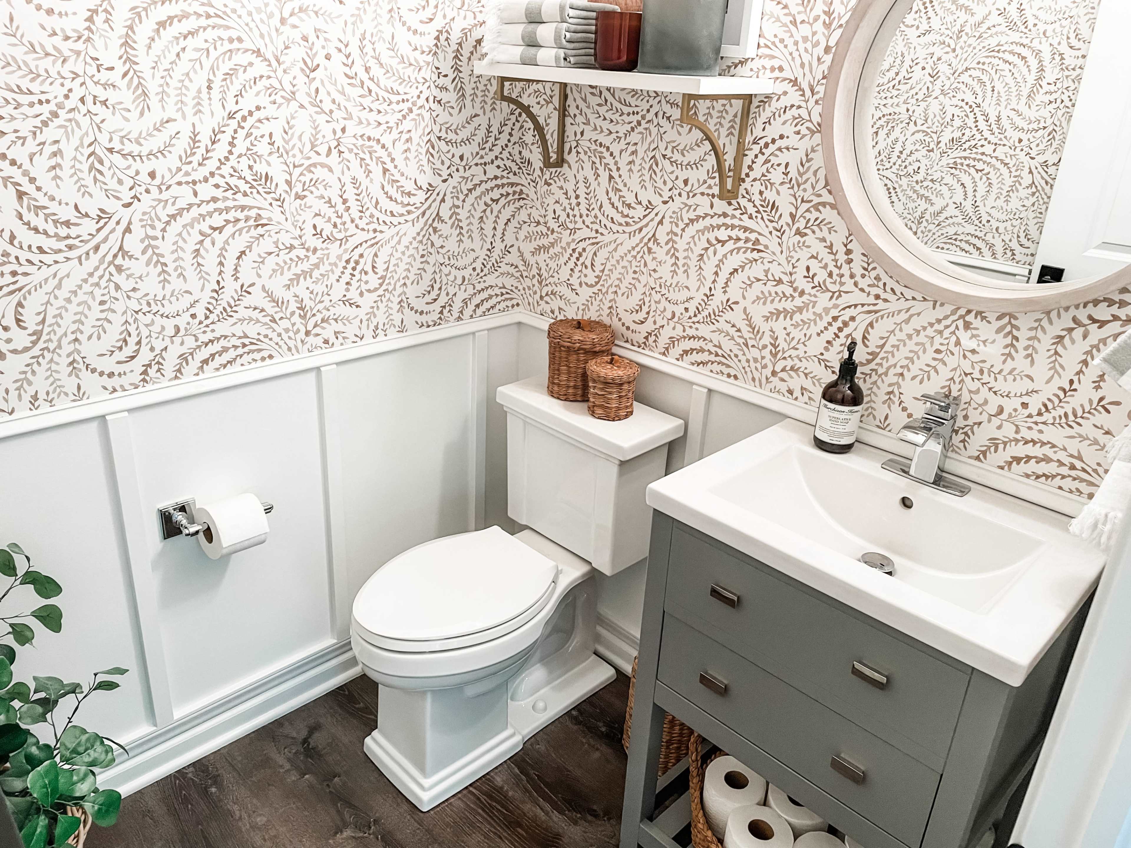 A small, stylish bathroom features a white toilet, a gray vanity with a sink, and a round mirror above, accented by patterned wallpaper and wicker storage baskets.