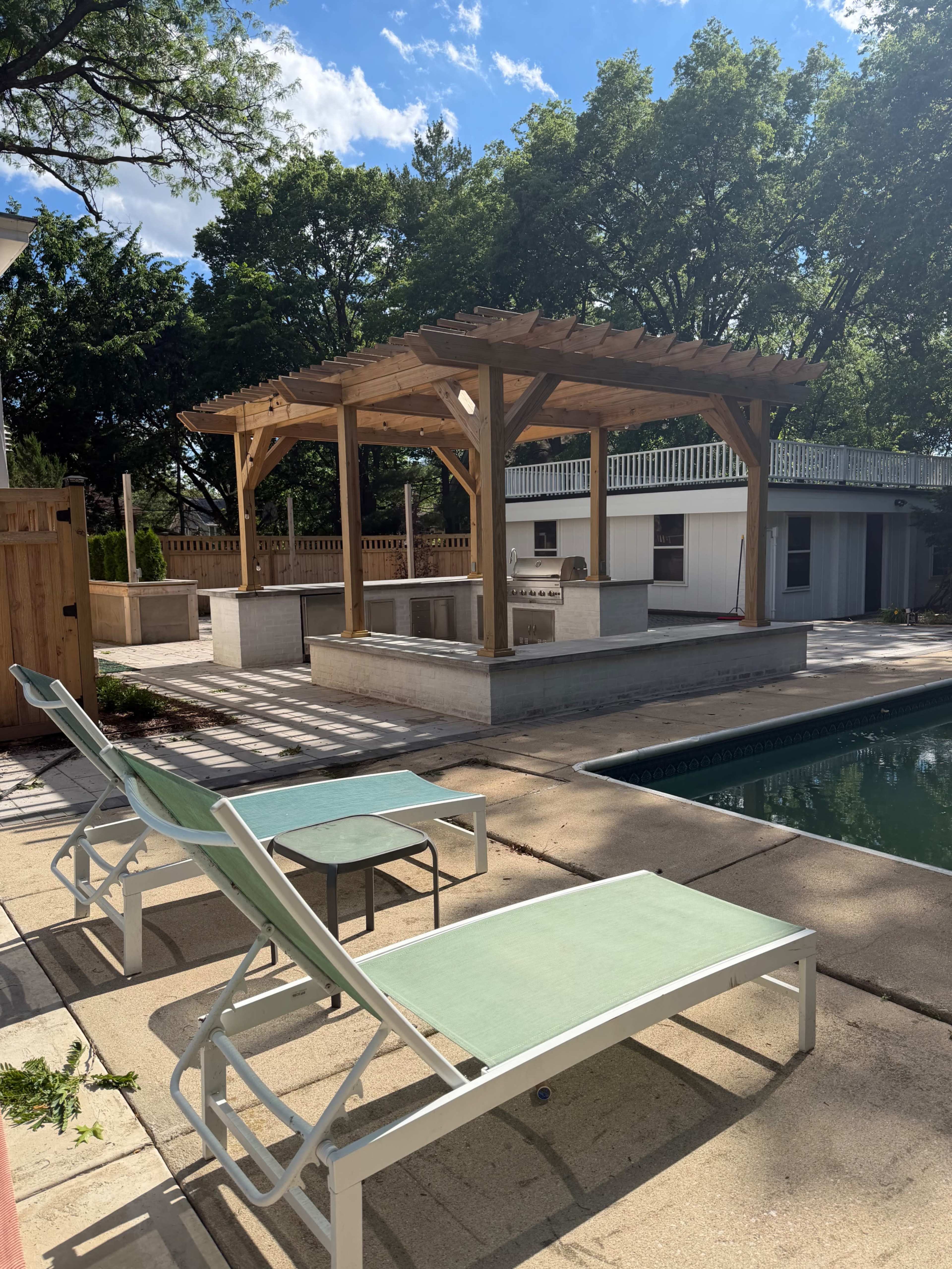 The image shows a backyard with a wooden pergola over outdoor kitchen amenities, a pool, and two green lounge chairs.