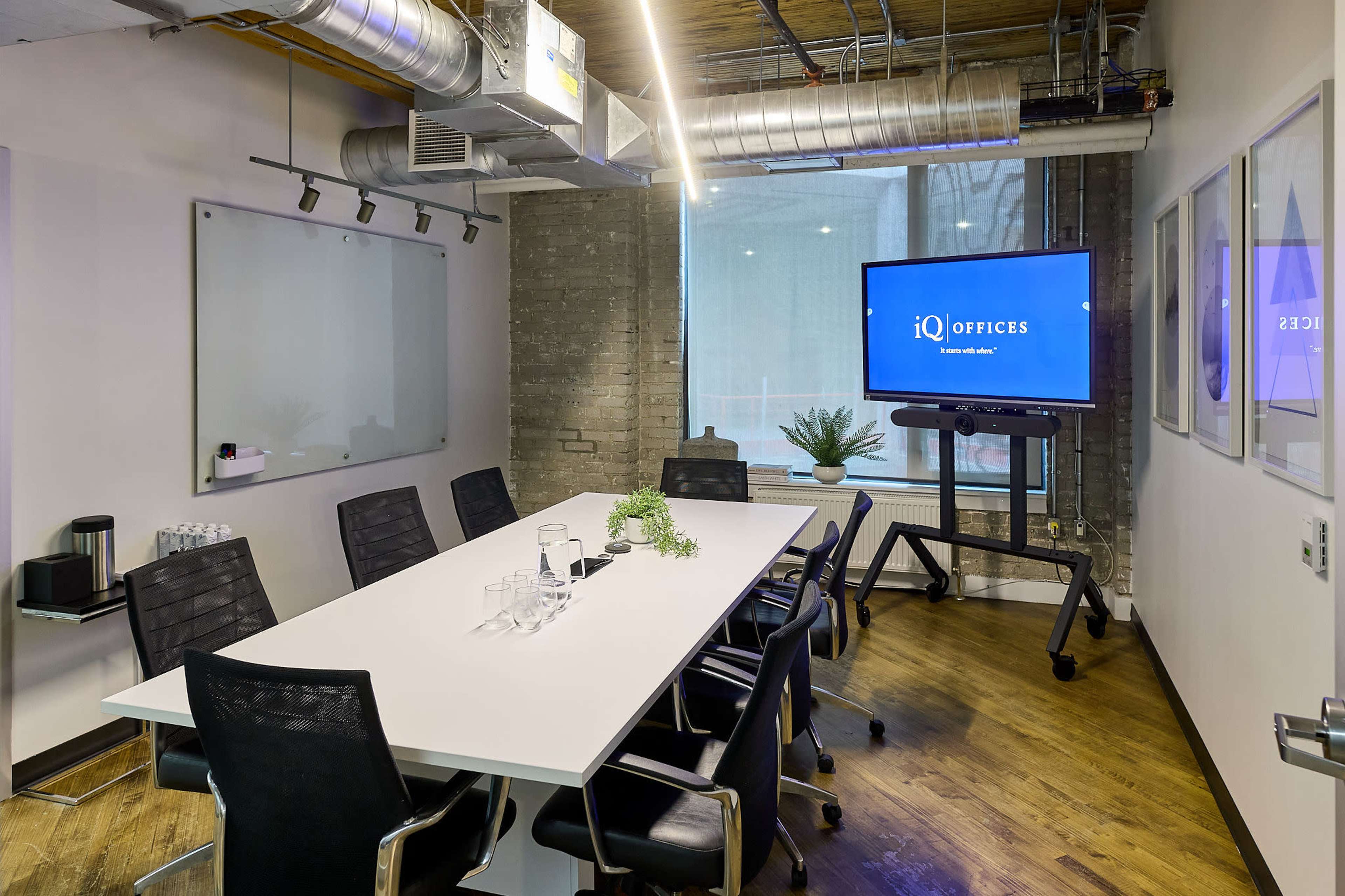 The image shows a modern conference room featuring a long white table surrounded by black chairs, a presentation screen, and large windows letting in natural light.