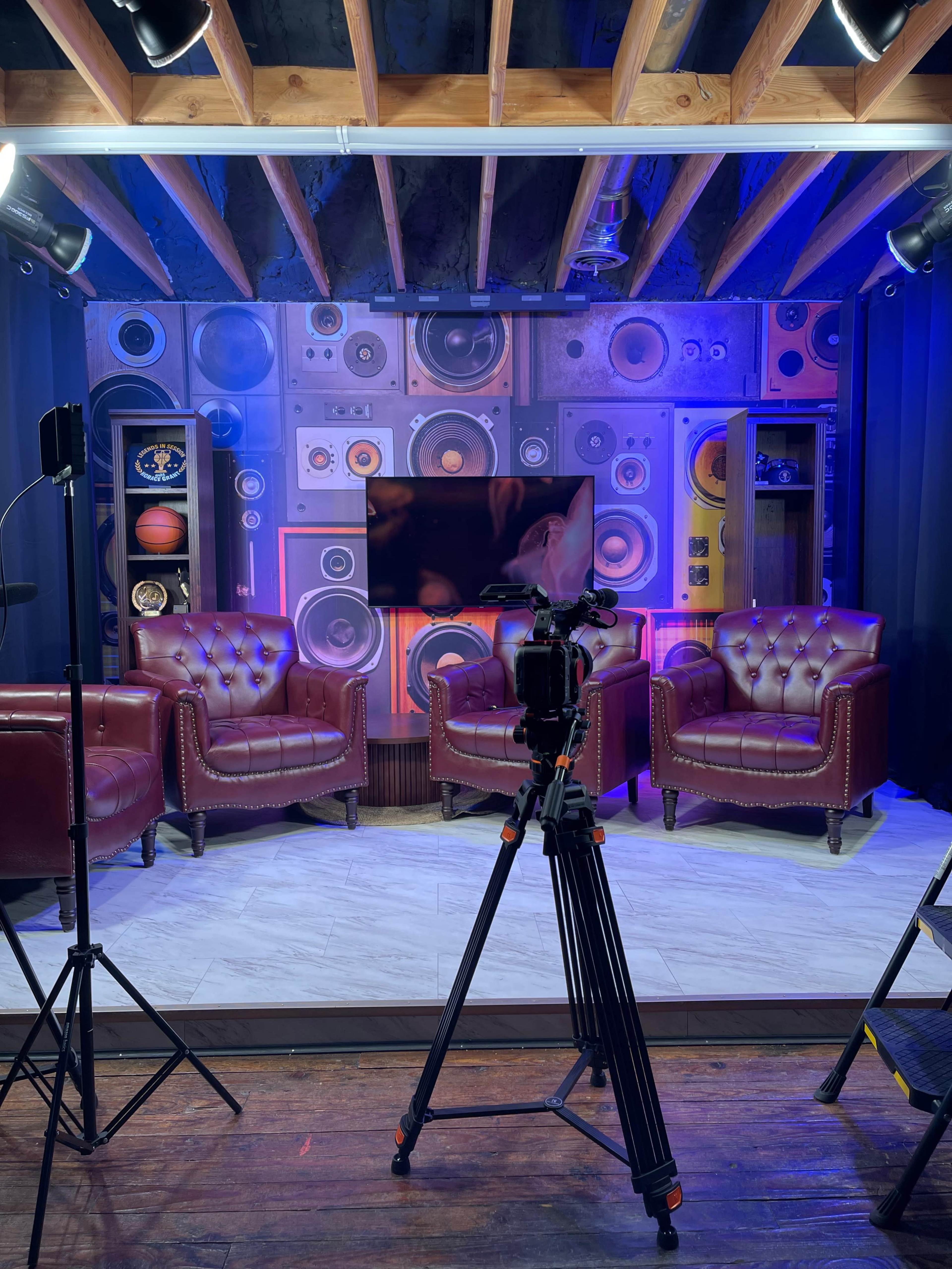 The image shows a television studio setup featuring two leather chairs, a large screen, and a backdrop adorned with vintage speakers and sports memorabilia, all under studio lighting.