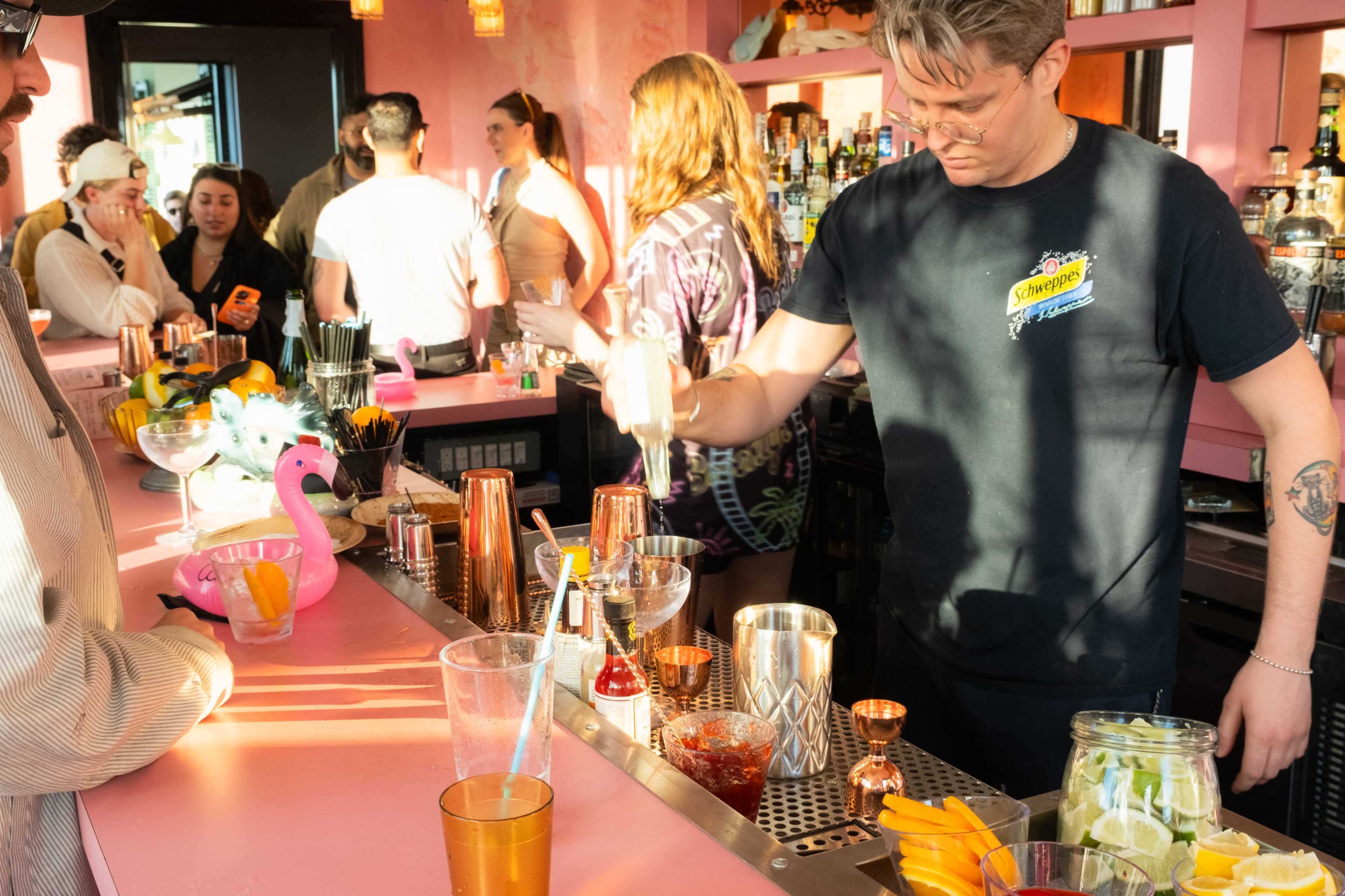 A bartender prepares cocktails at a vibrant bar filled with patrons in a pink-themed setting.
