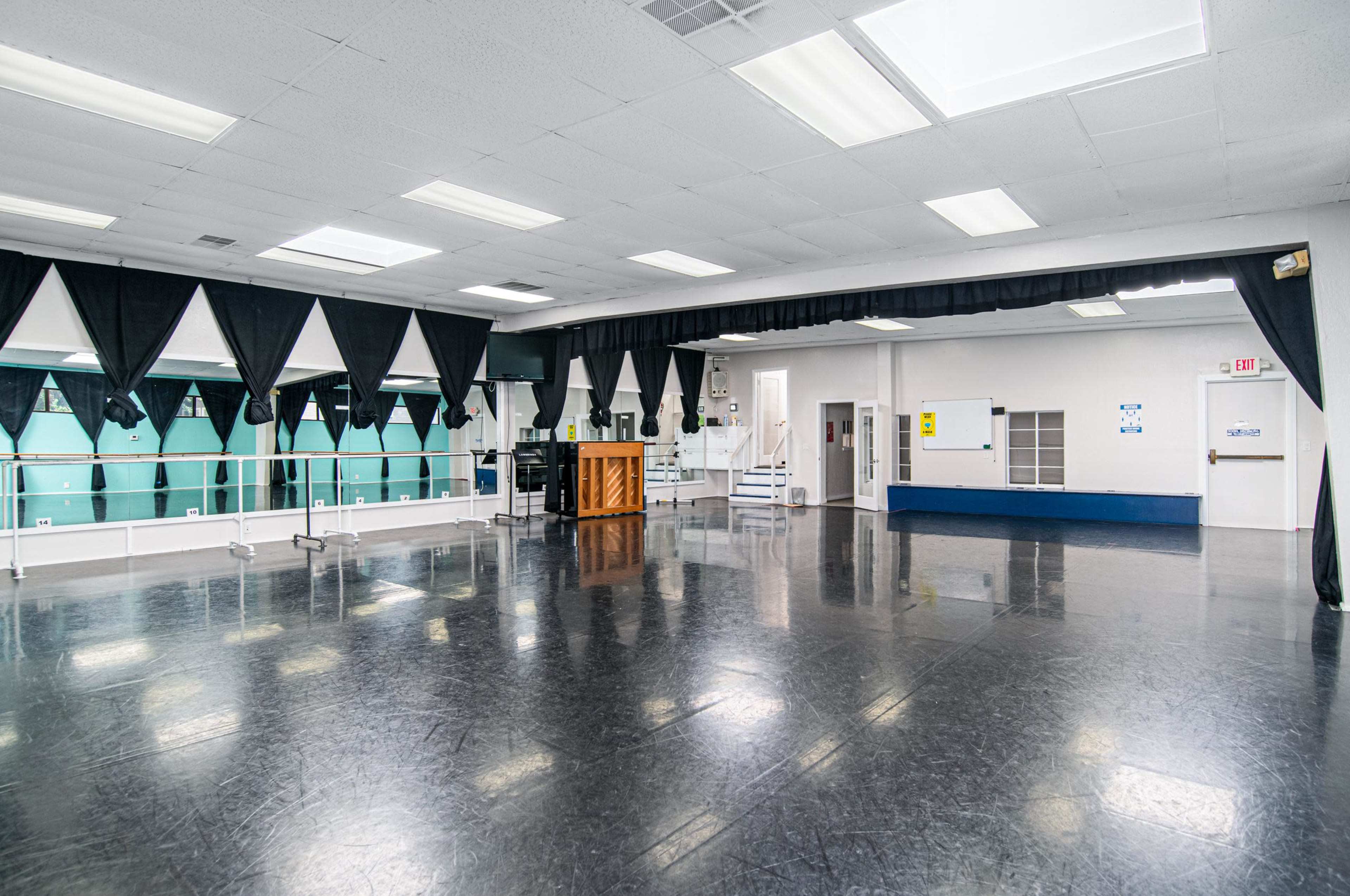 A spacious dance studio with a black floor, large mirrors, and a grand piano, surrounded by curtain-draped windows.