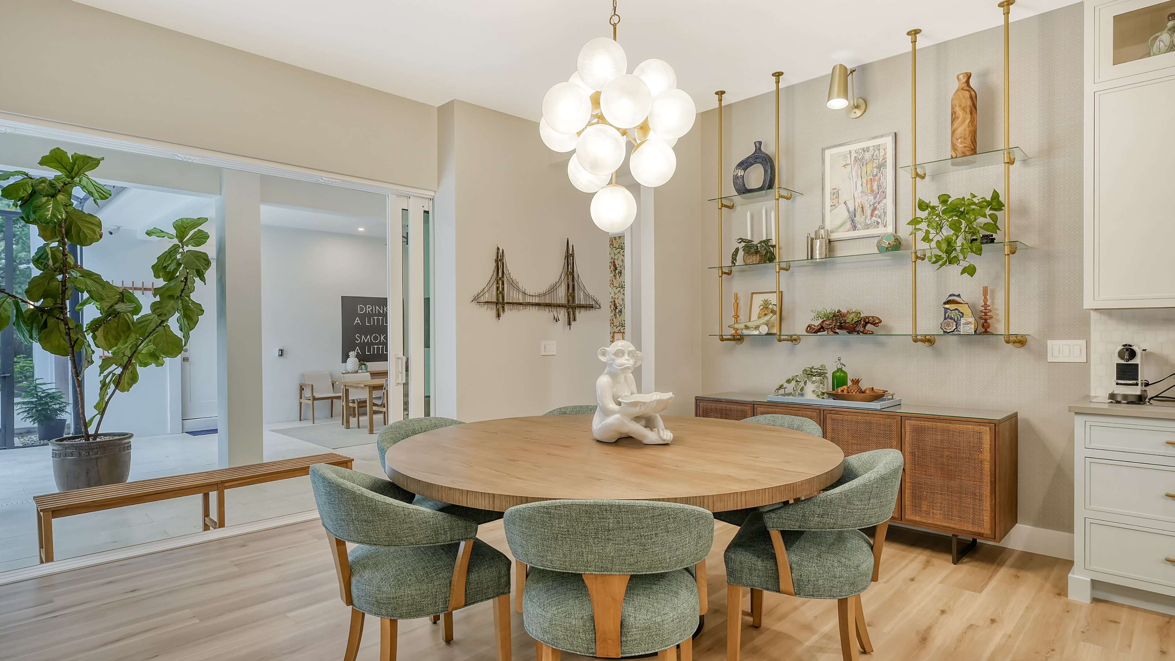 A dining area features a round wooden table surrounded by upholstered chairs, with a decorative sculpture on the table and various plants and artwork displayed on shelves.
