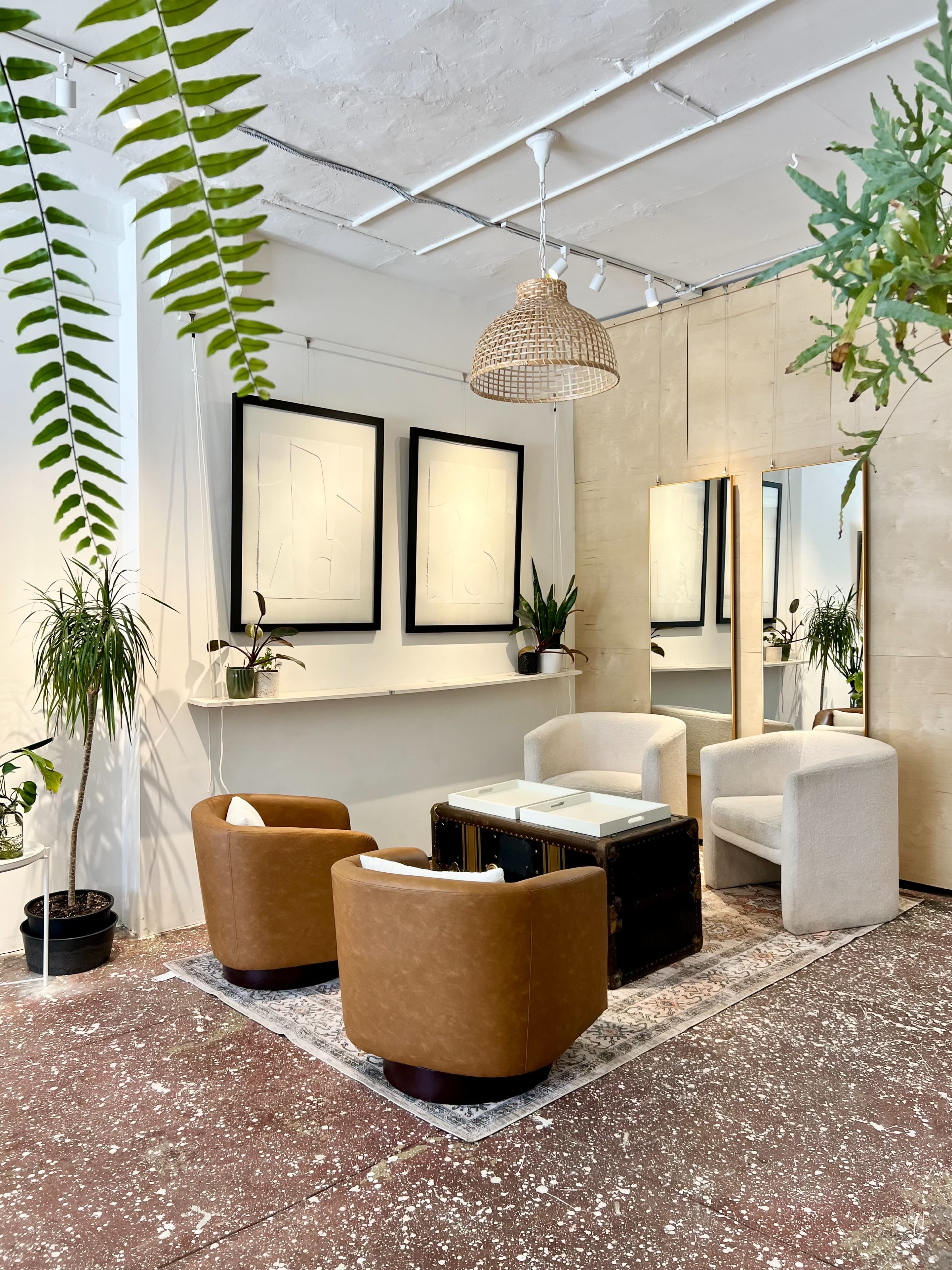 The image shows a modern interior space featuring two brown armchairs and two white chairs around a coffee table, with plants, artwork on the walls, and a large mirror.