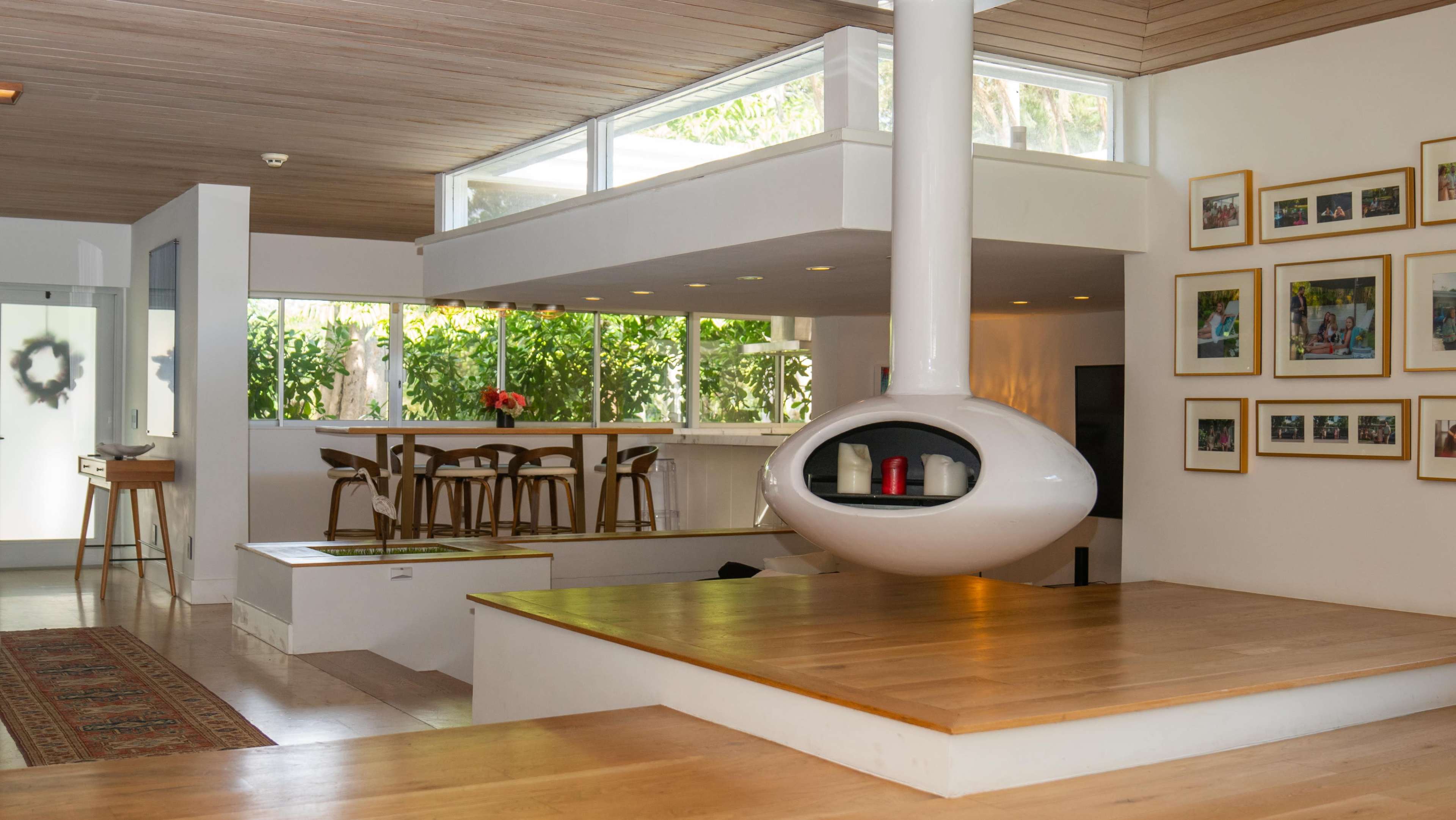 The interior of a modern house features an open living space with a distinctive white cylindrical fireplace, wooden flooring, and a dining area with bar stools.