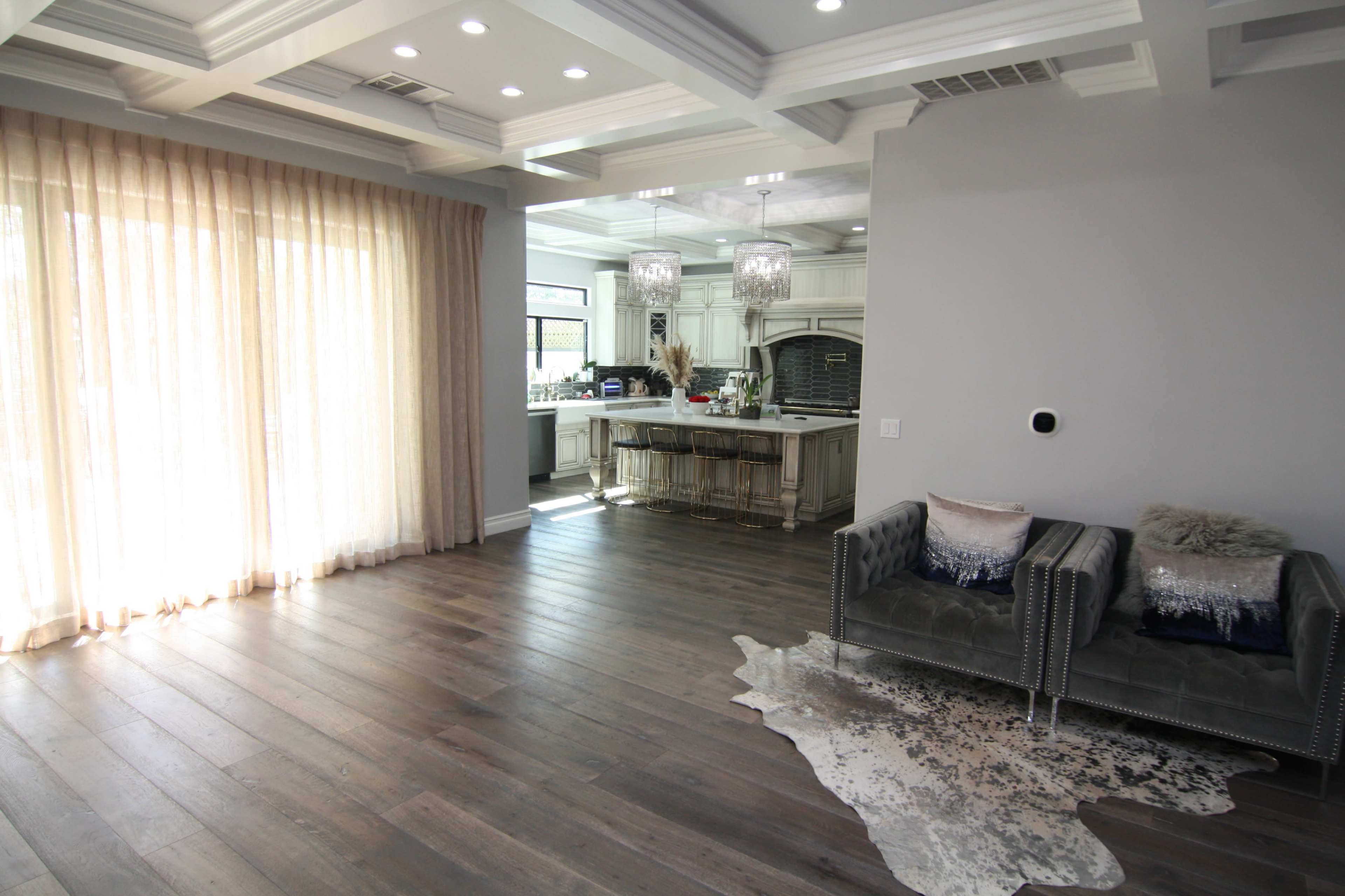 A spacious living area features hardwood floors, large windows with sheer curtains, and a view of an adjacent kitchen with an island and pendant lighting.