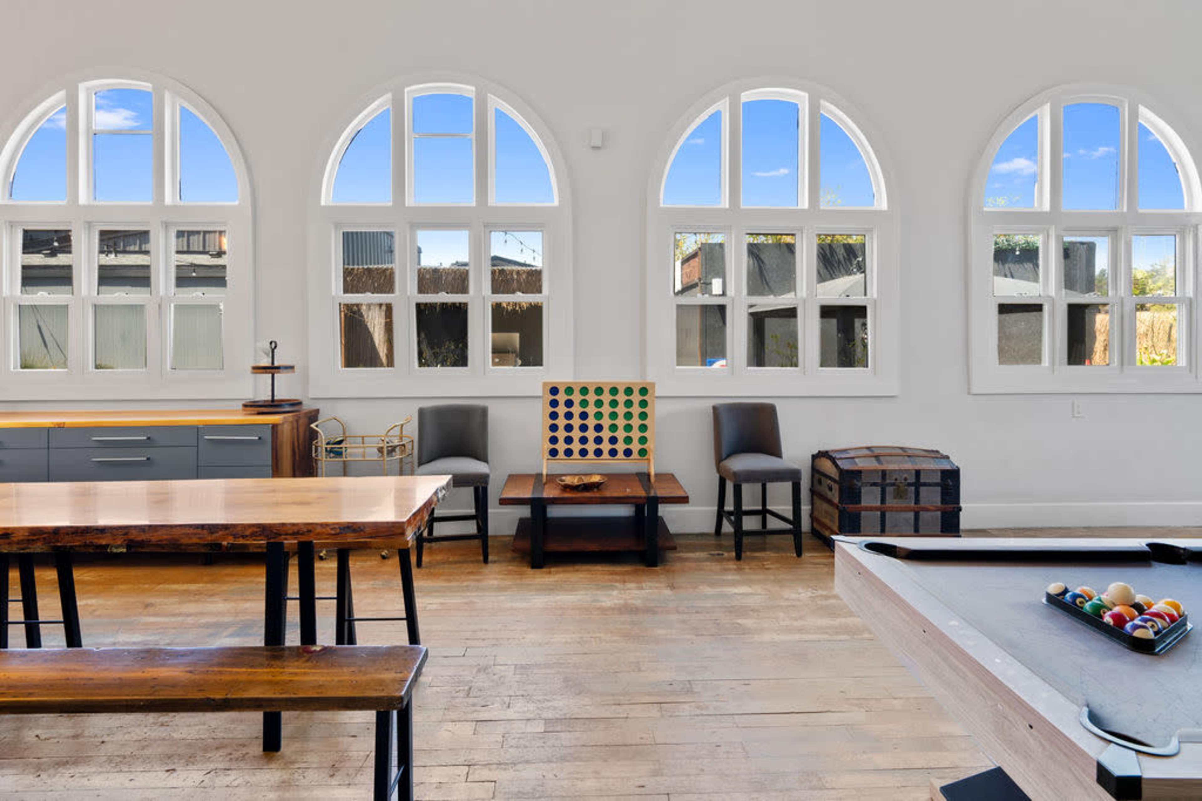 The interior features a spacious room with large arched windows, wooden tables, and a seating area, showcasing a mix of functional furniture and decor items.