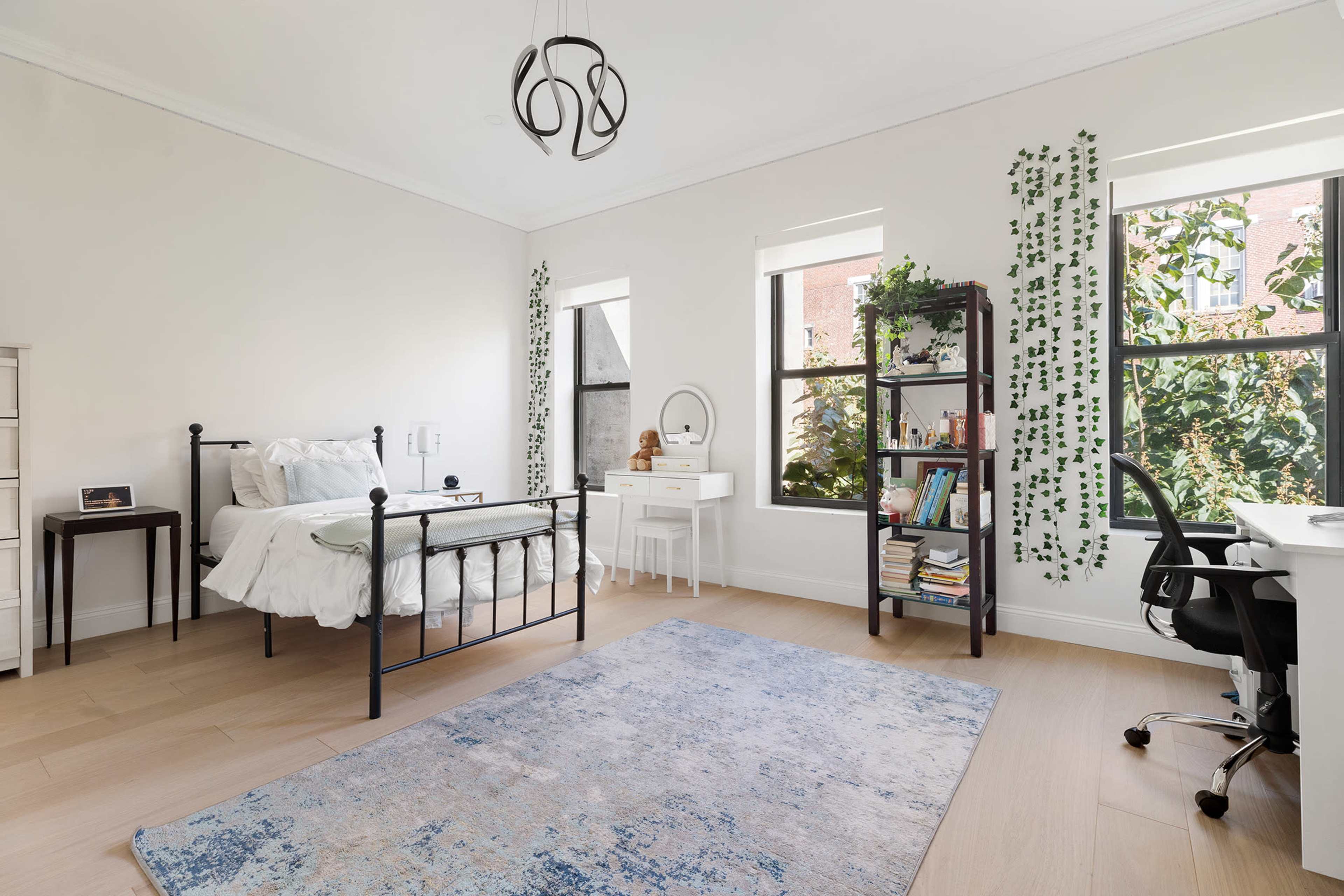 The image shows a bright, minimalist bedroom featuring a single metal bed, a white desk with a mirror, a bookshelf filled with books, and large windows with greenery visible outside.
