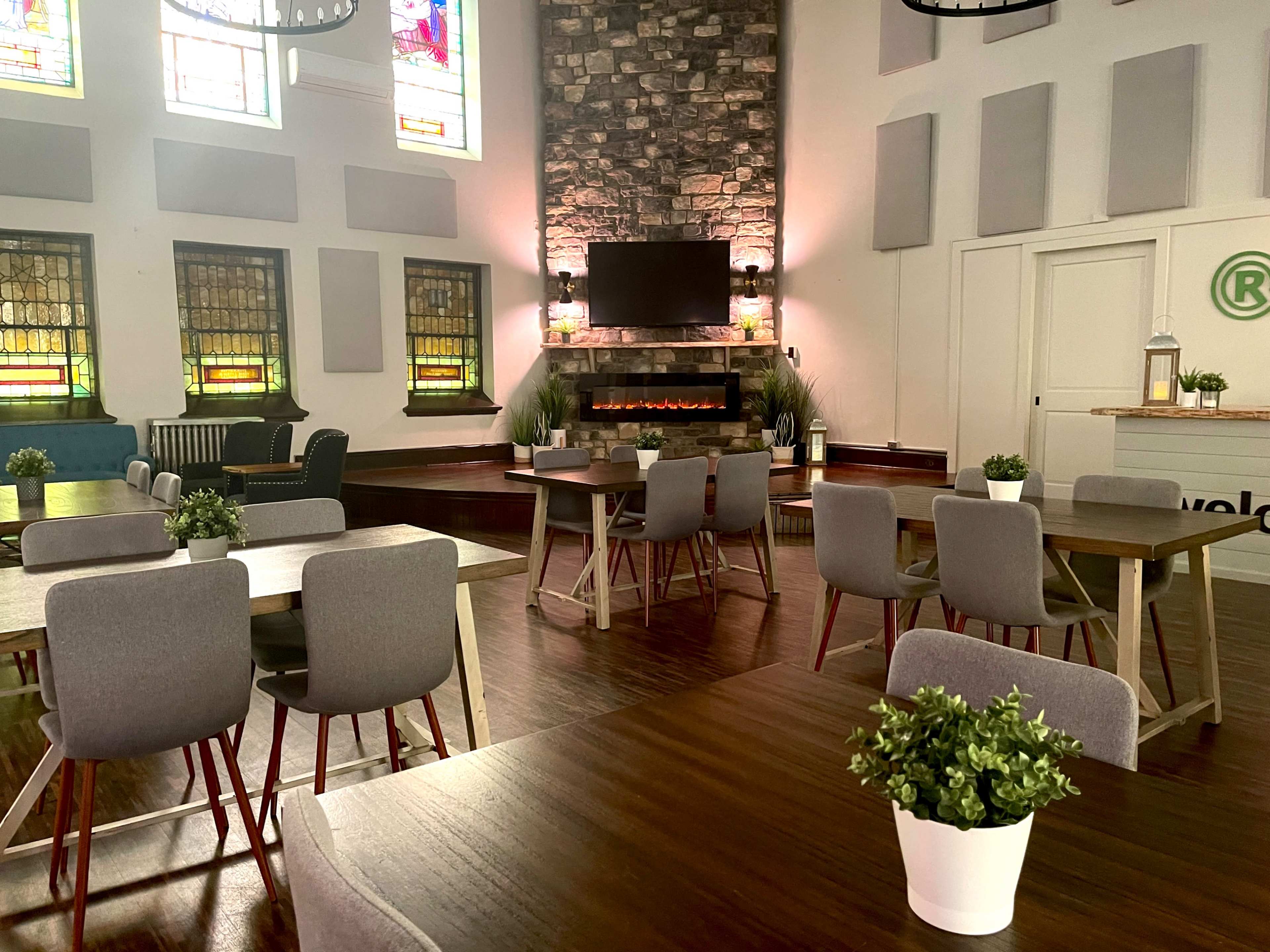 A spacious, well-lit room with multiple tables arranged around a modern fireplace and stained glass windows.