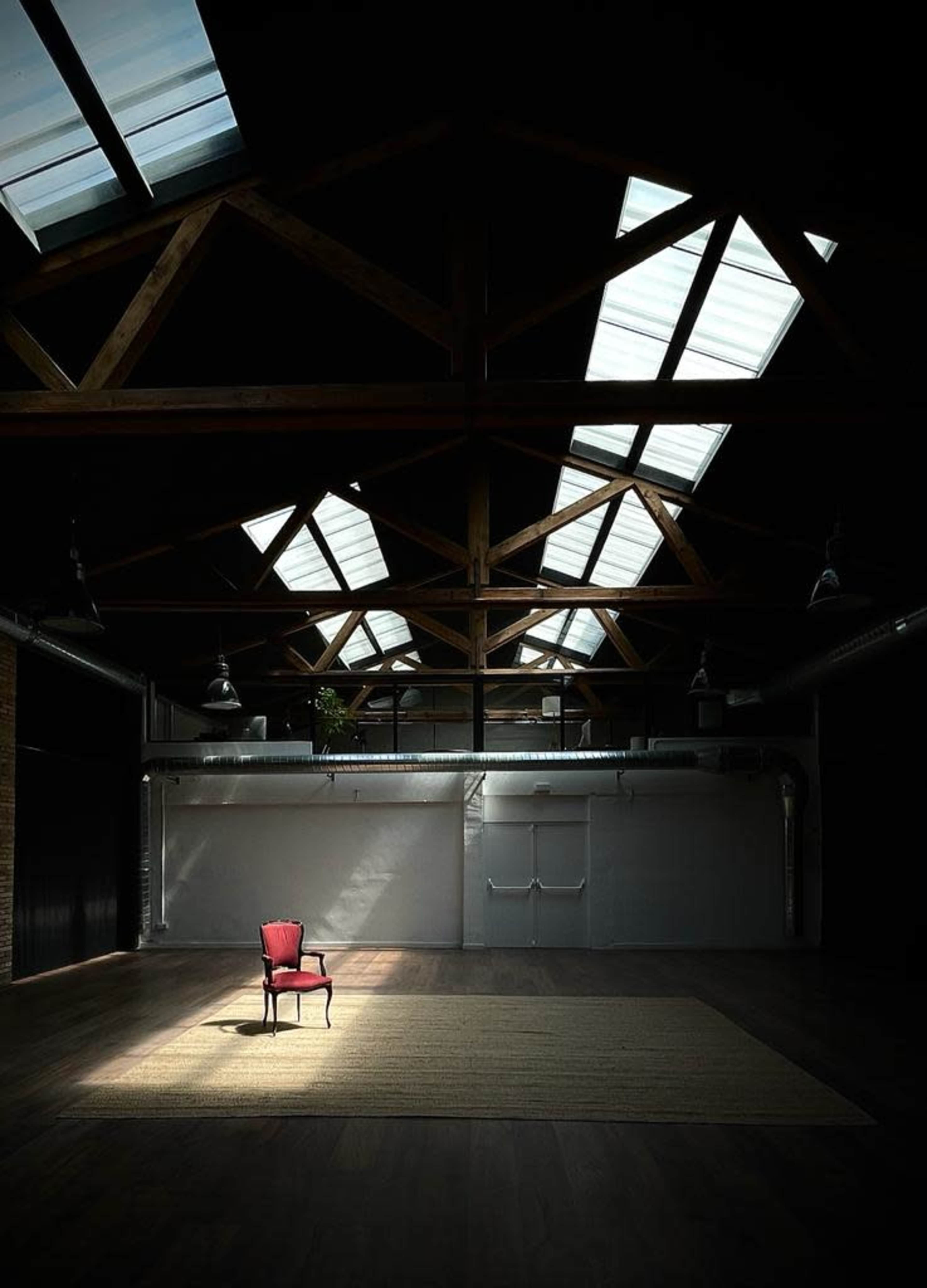 A red chair is positioned on a large rug in a spacious, dimly lit room with high, wooden beams and skylights.