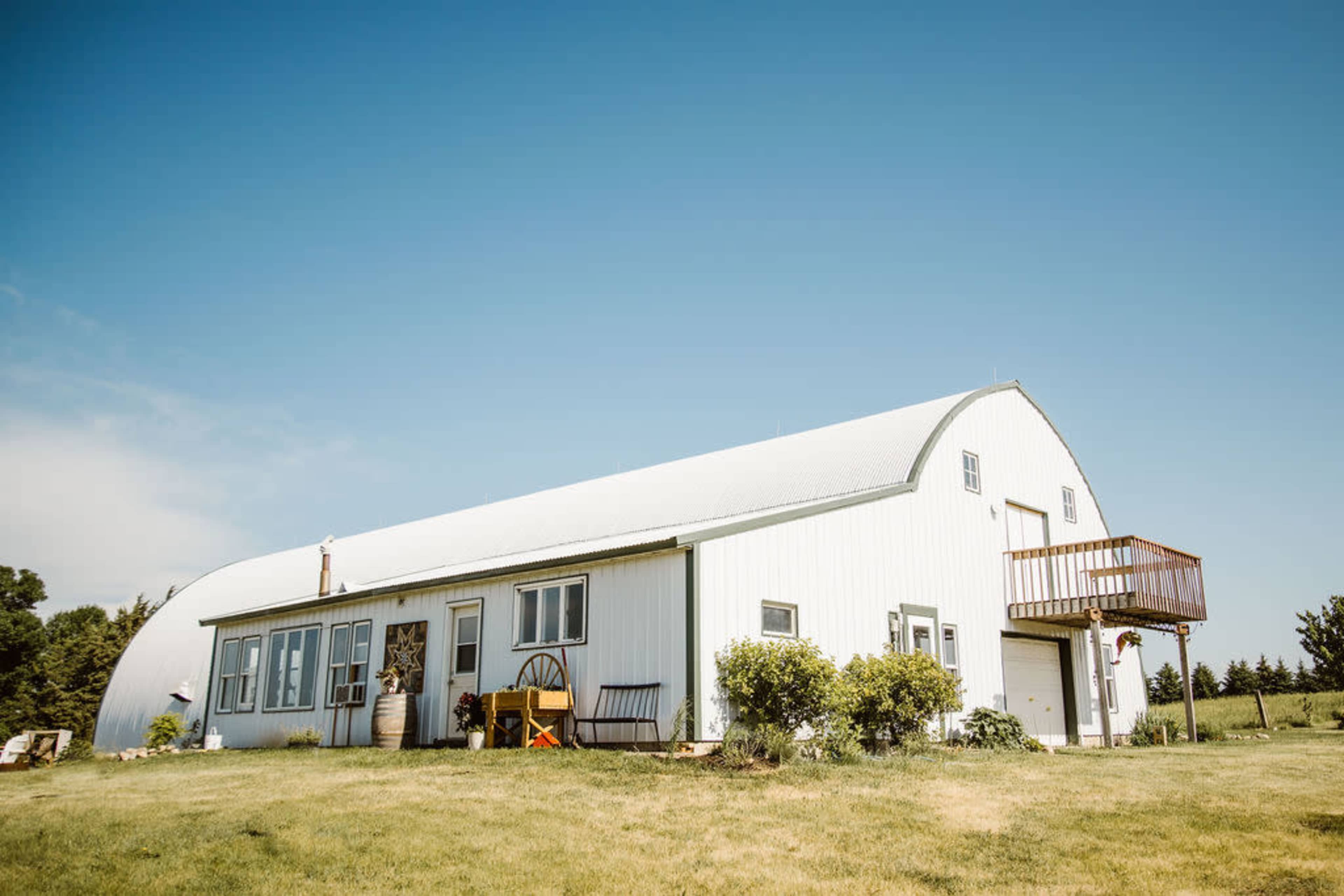 A large, white barn with a curved roof and a wooden deck is situated on a grassy field under a clear blue sky.