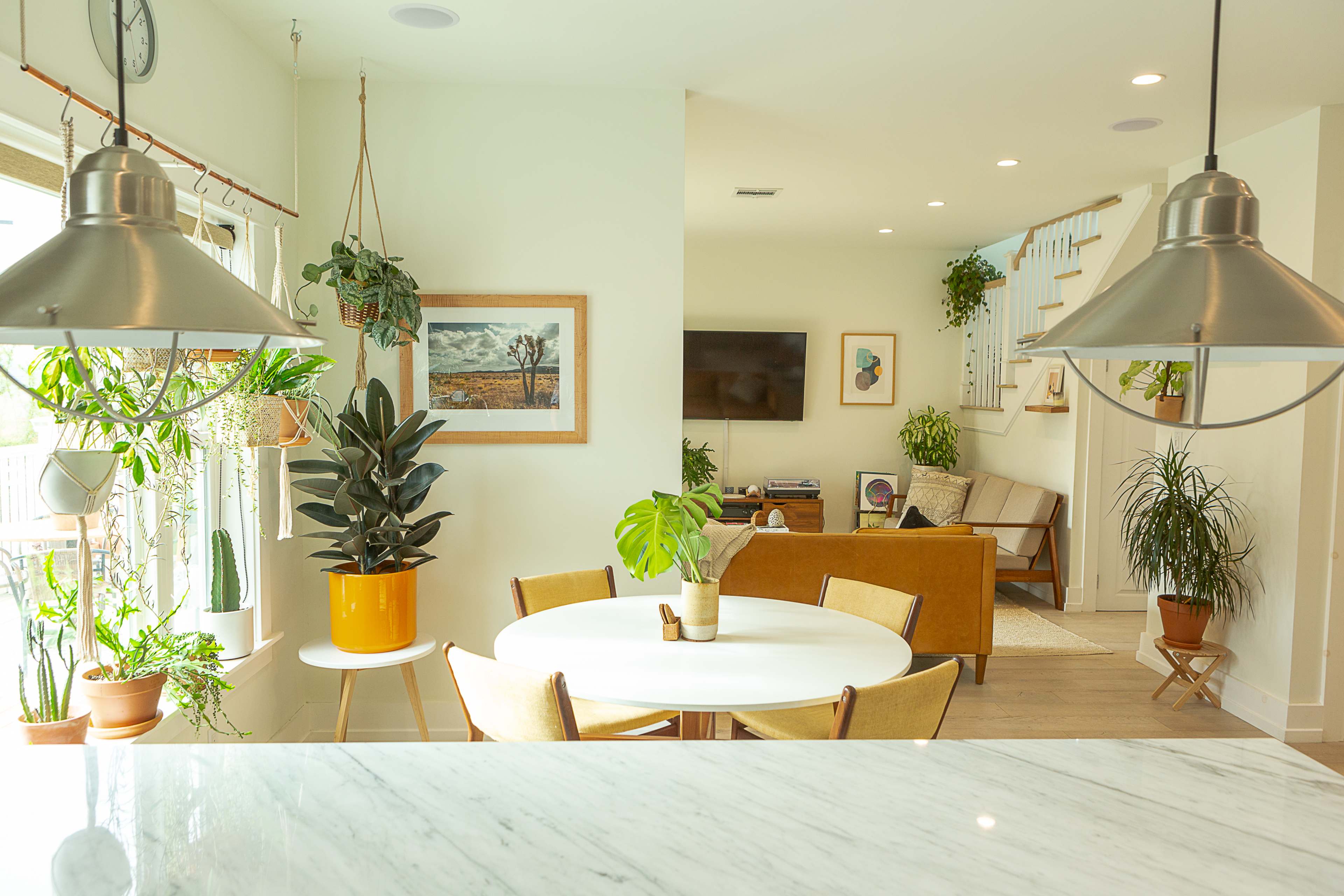 A bright, modern living space features a round dining table surrounded by chairs, with plants adorning the windows and a cozy seating area visible in the background.