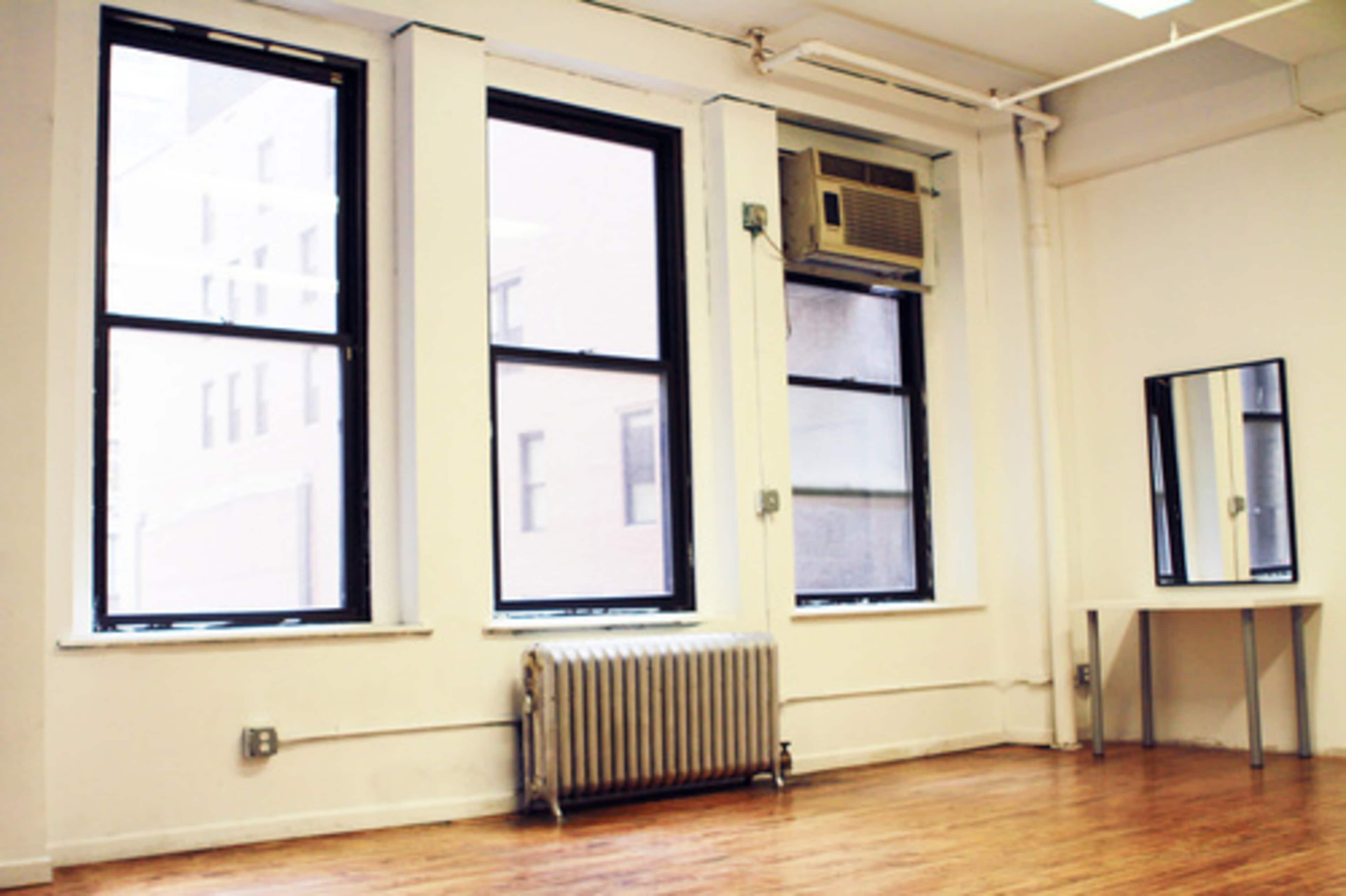 A room with three large windows, an air conditioning unit, a radiator, and a small table with a mirror.