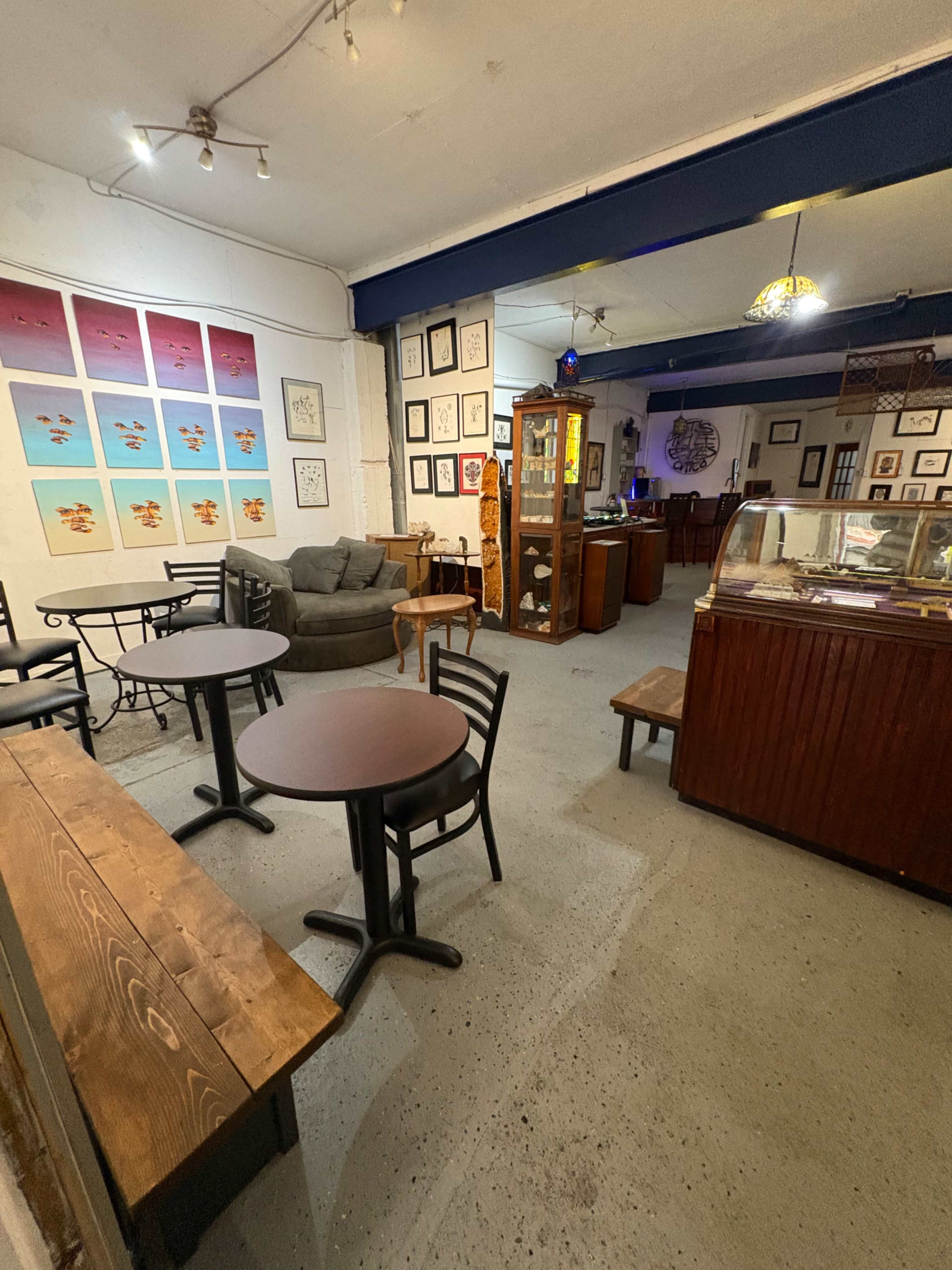 The image shows a cozy interior of a café with two small tables, a couch, framed artwork on the walls, and a display case.