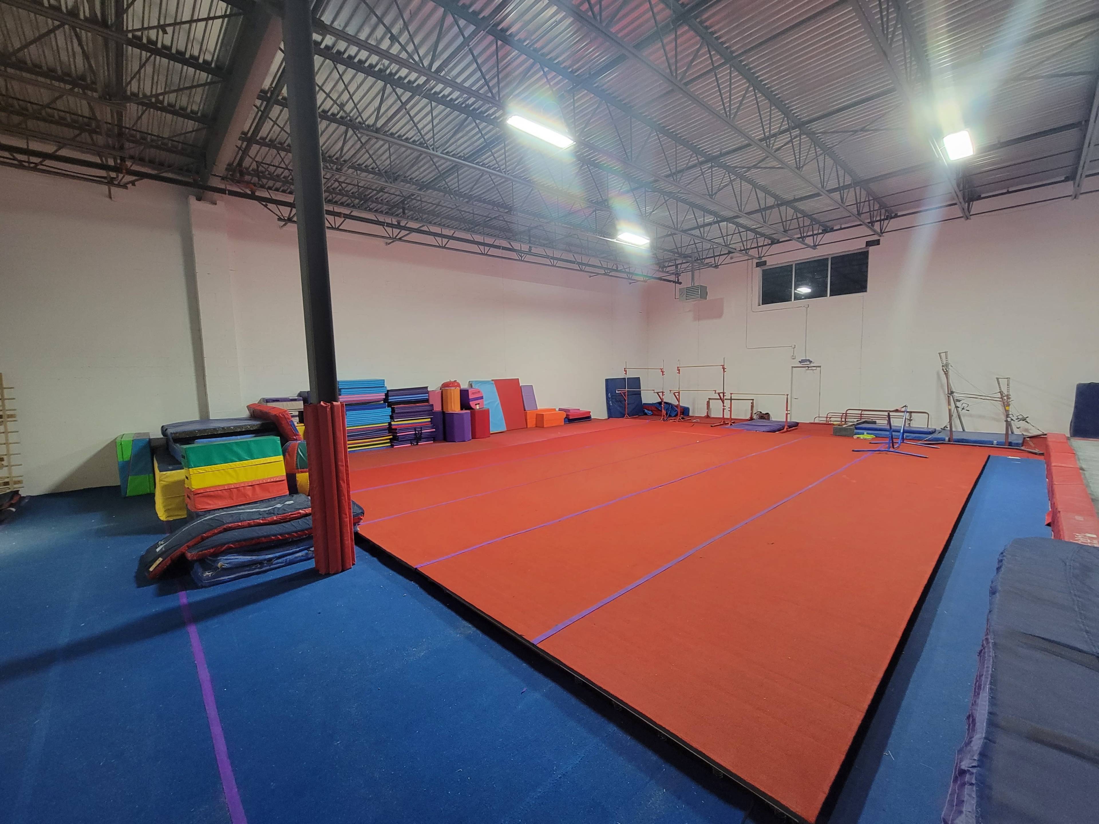 An empty gymnastics facility with red and blue mats arranged on the floor and various gymnastic equipment positioned along the walls.