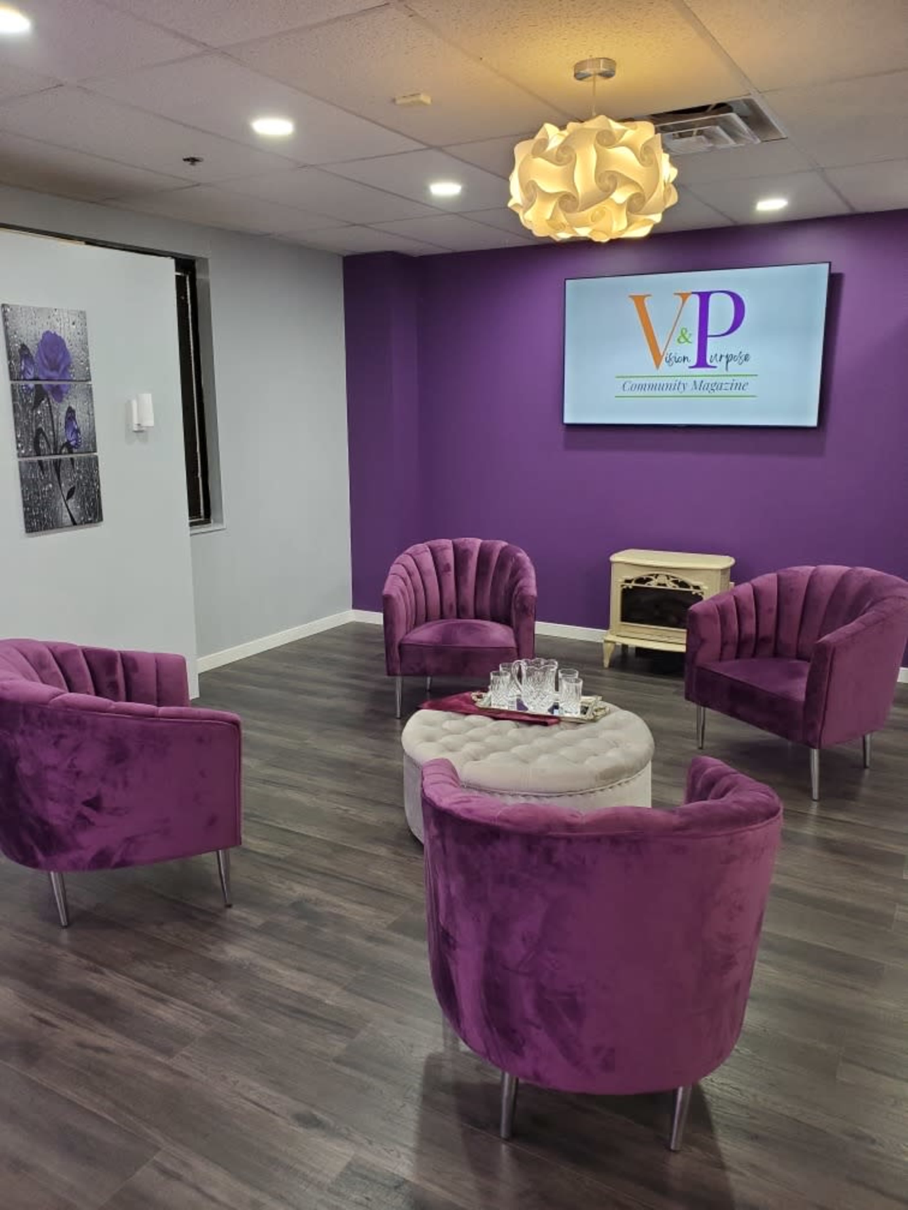 A modern waiting area featuring four purple velvet chairs arranged around a small circular table in front of a wall-mounted screen.