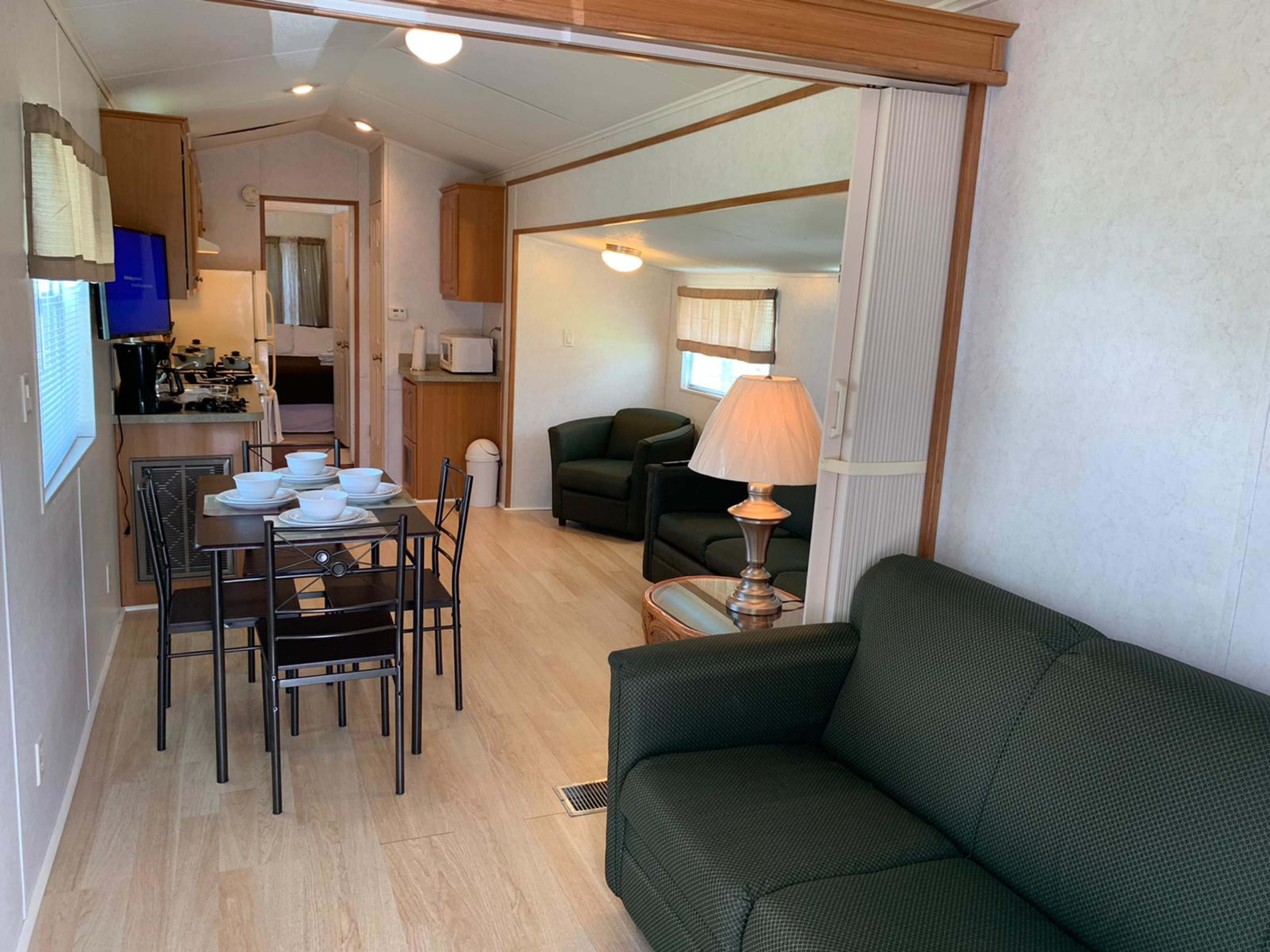 A dining area with a table set for six is positioned next to a living space featuring a green couch and a lamp, with a kitchen visible in the background.