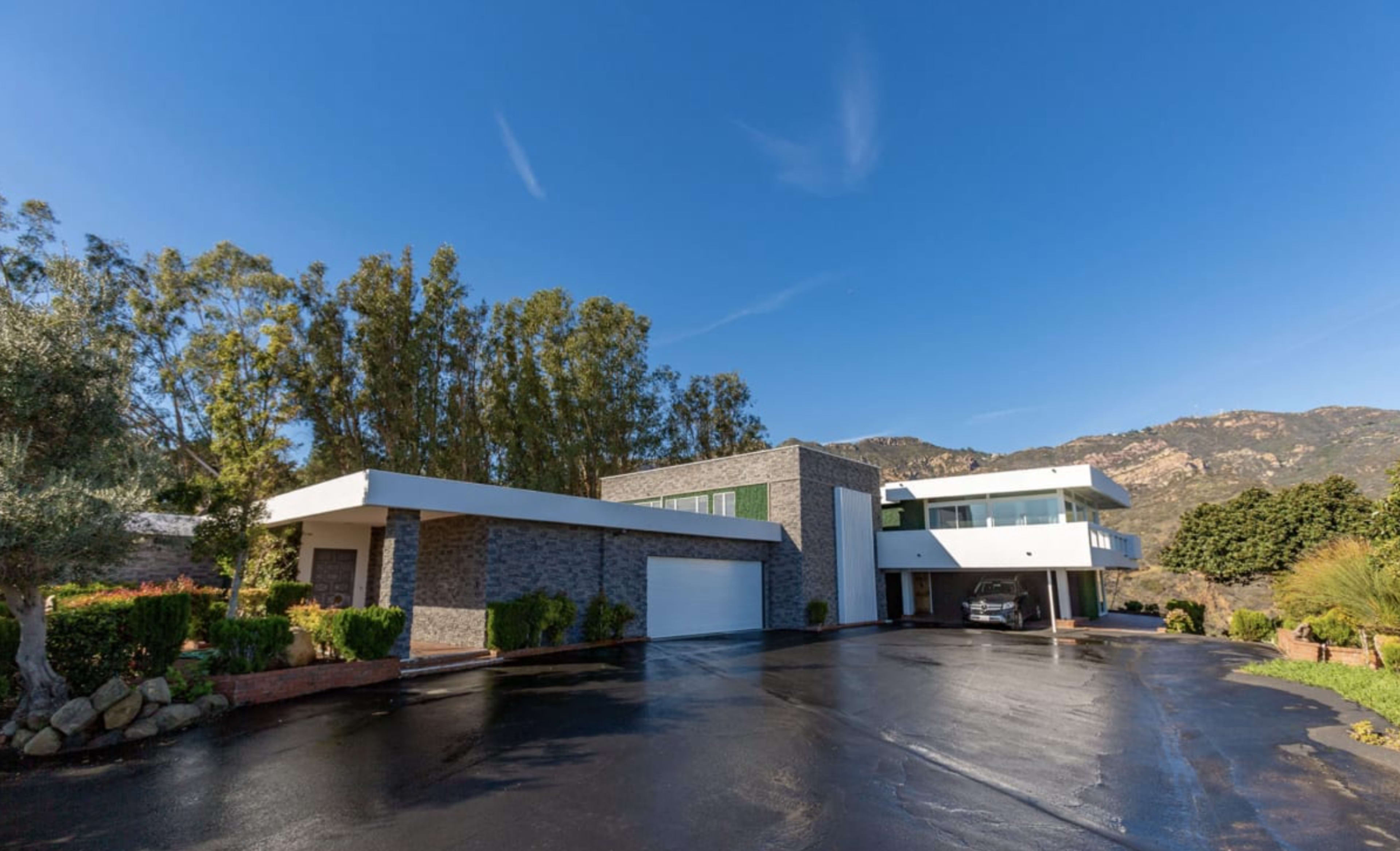 A modern two-story house with a stone facade and a circular driveway surrounded by greenery and mountains in the background.