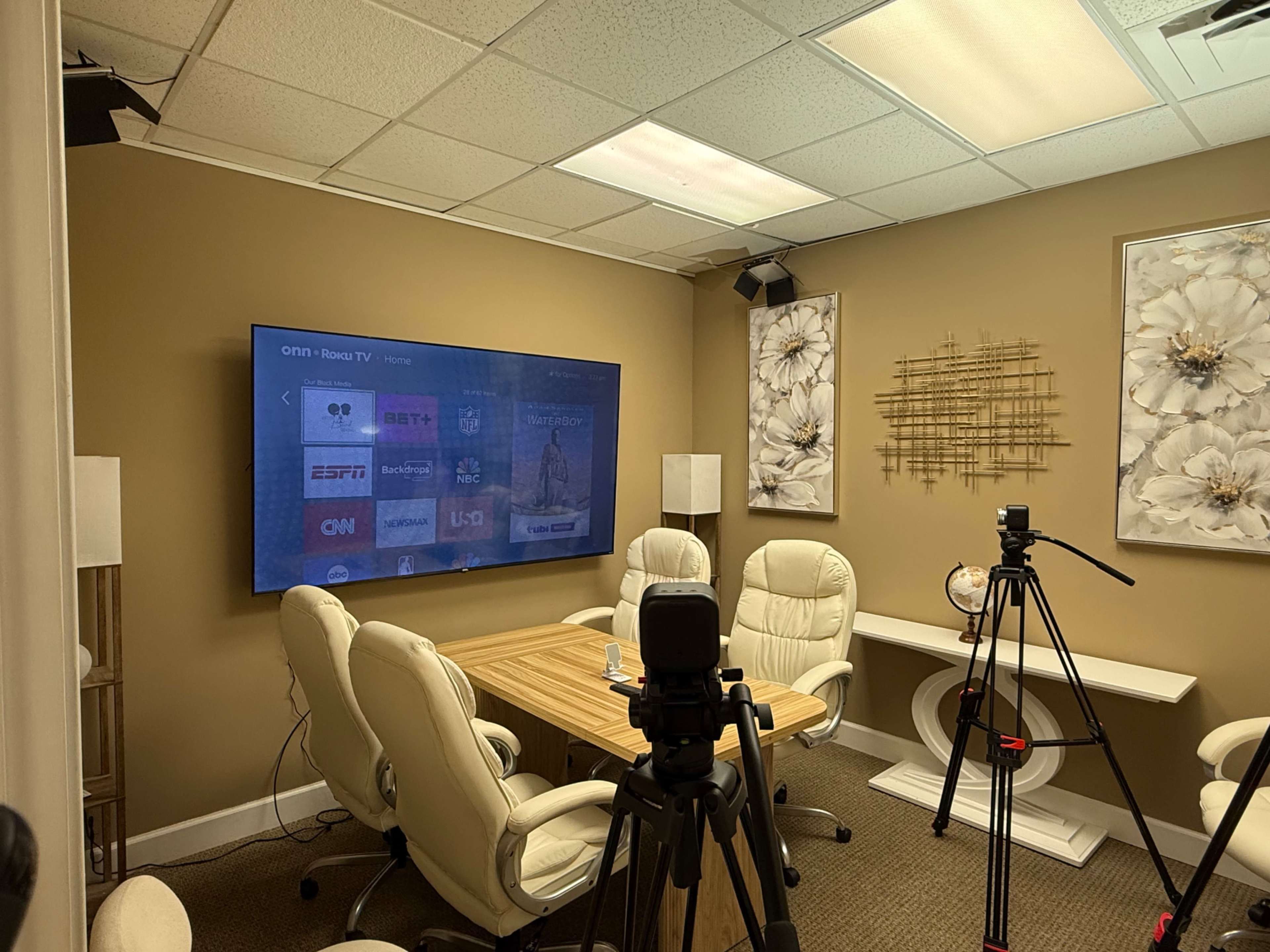 The image depicts a conference room with a large screen displaying various TV channels, surrounded by a wooden table and white chairs, with camera equipment set up for recording.