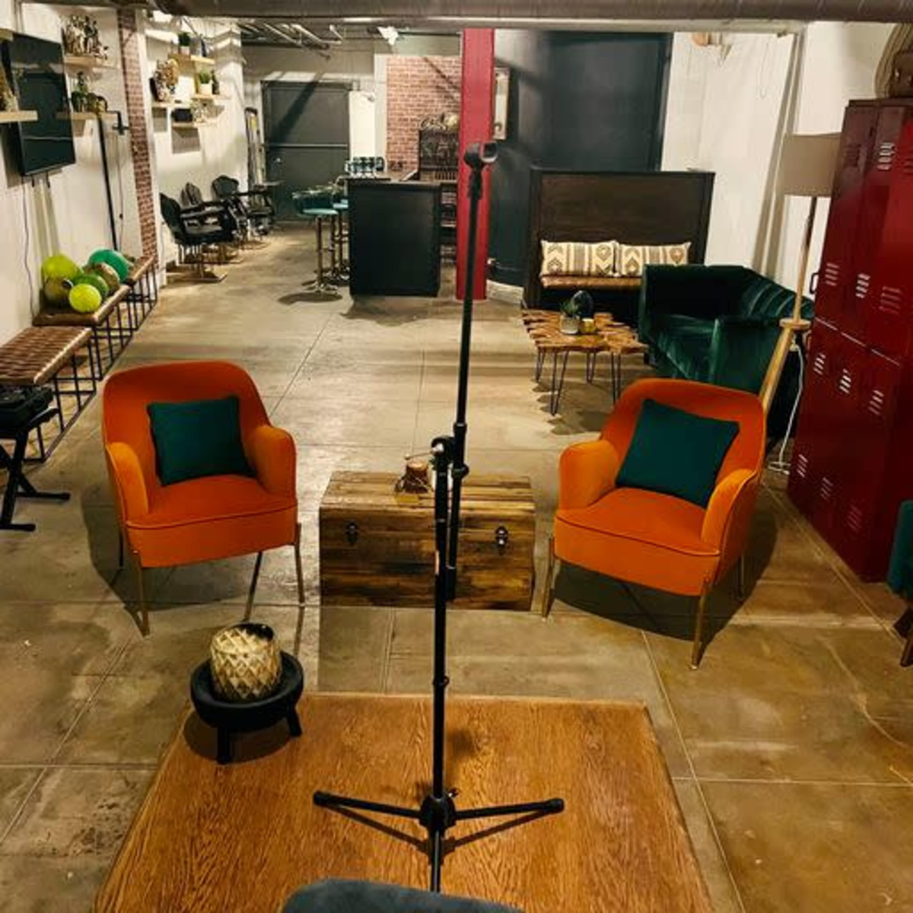 A cozy lounge area featuring two orange chairs, a wooden table, and a microphone stand in an industrial-style space.