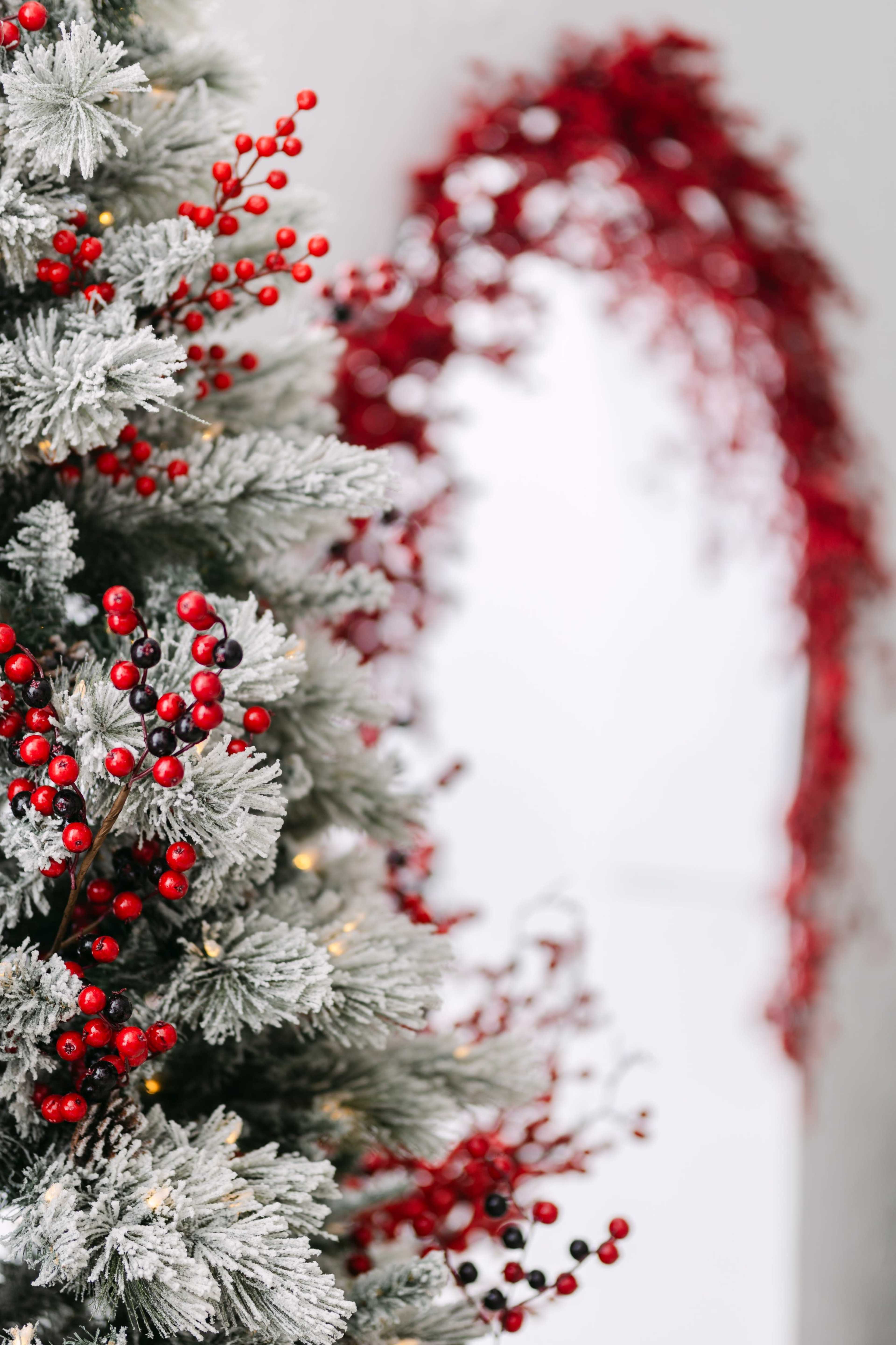 A frosted Christmas tree adorned with red berries and lights stands in front of a background featuring a garland of red decorations.