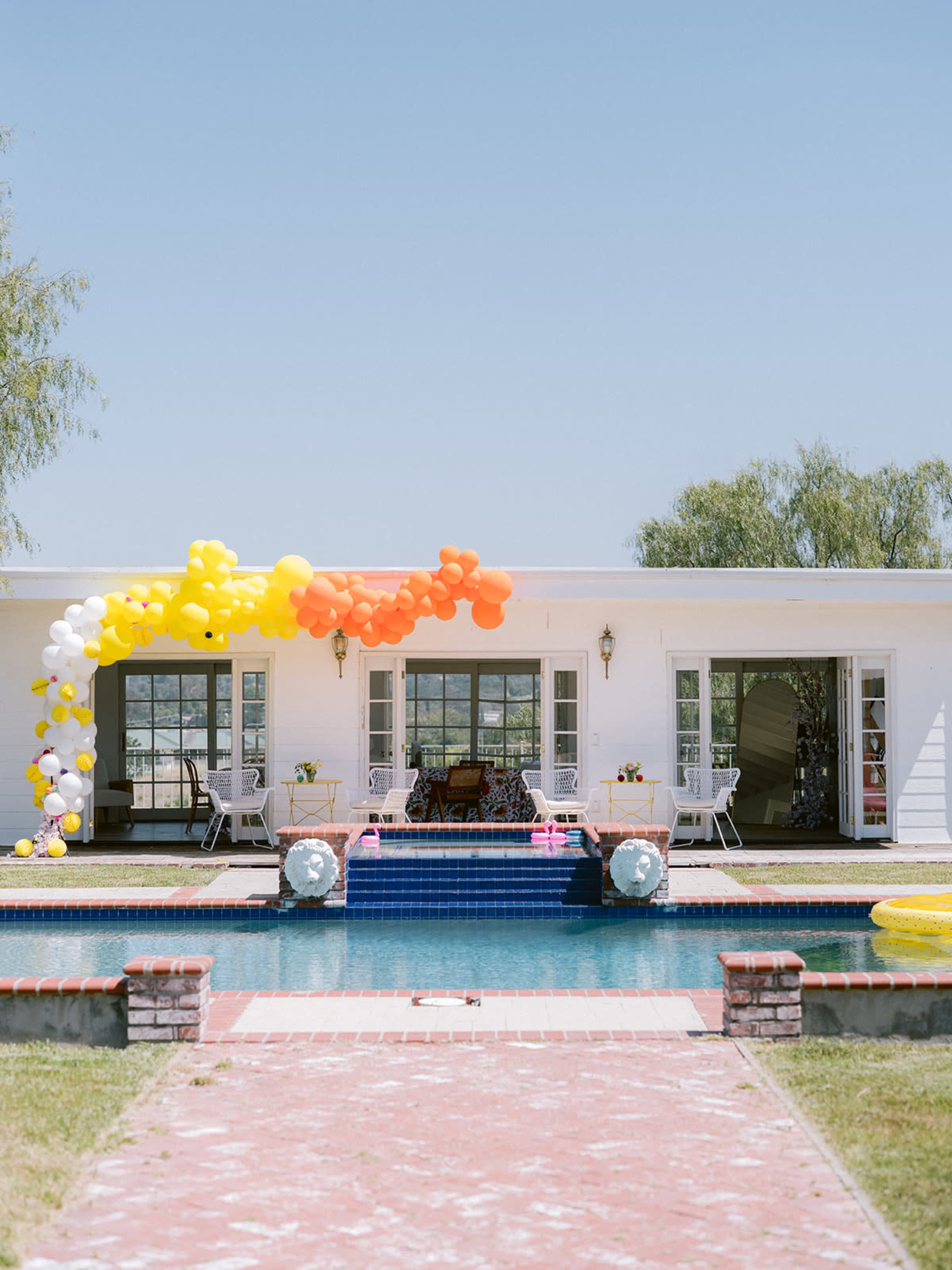 A colorful balloon arch stretches over a pool area in front of a white house with large windows and outdoor seating.