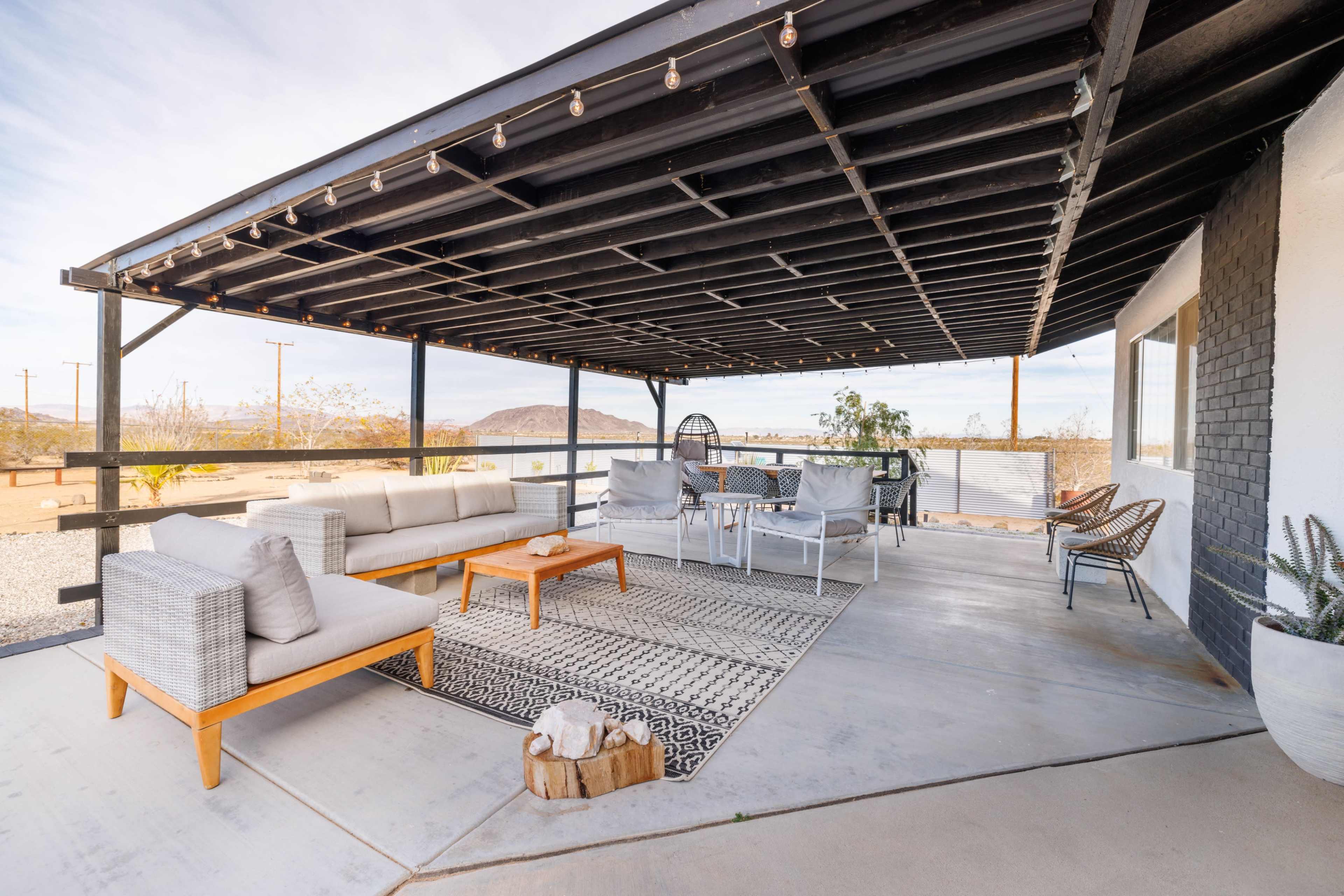A covered patio features a seating area with a gray sofa and chairs, a wooden coffee table, and decorative elements overlooking a desert landscape.