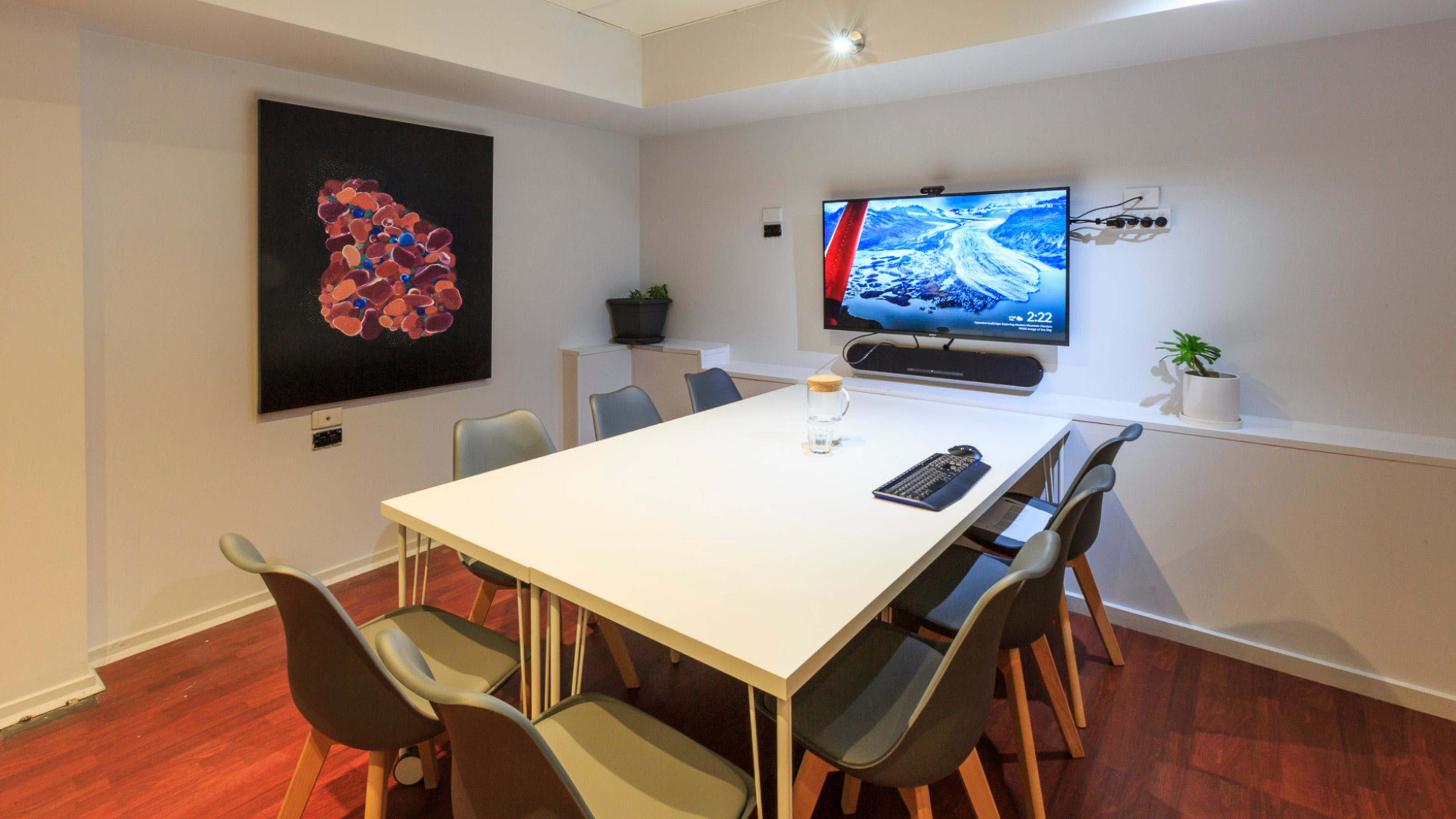 A modern conference room features a large white table surrounded by chairs, a wall-mounted television, and an artwork on the adjacent wall.
