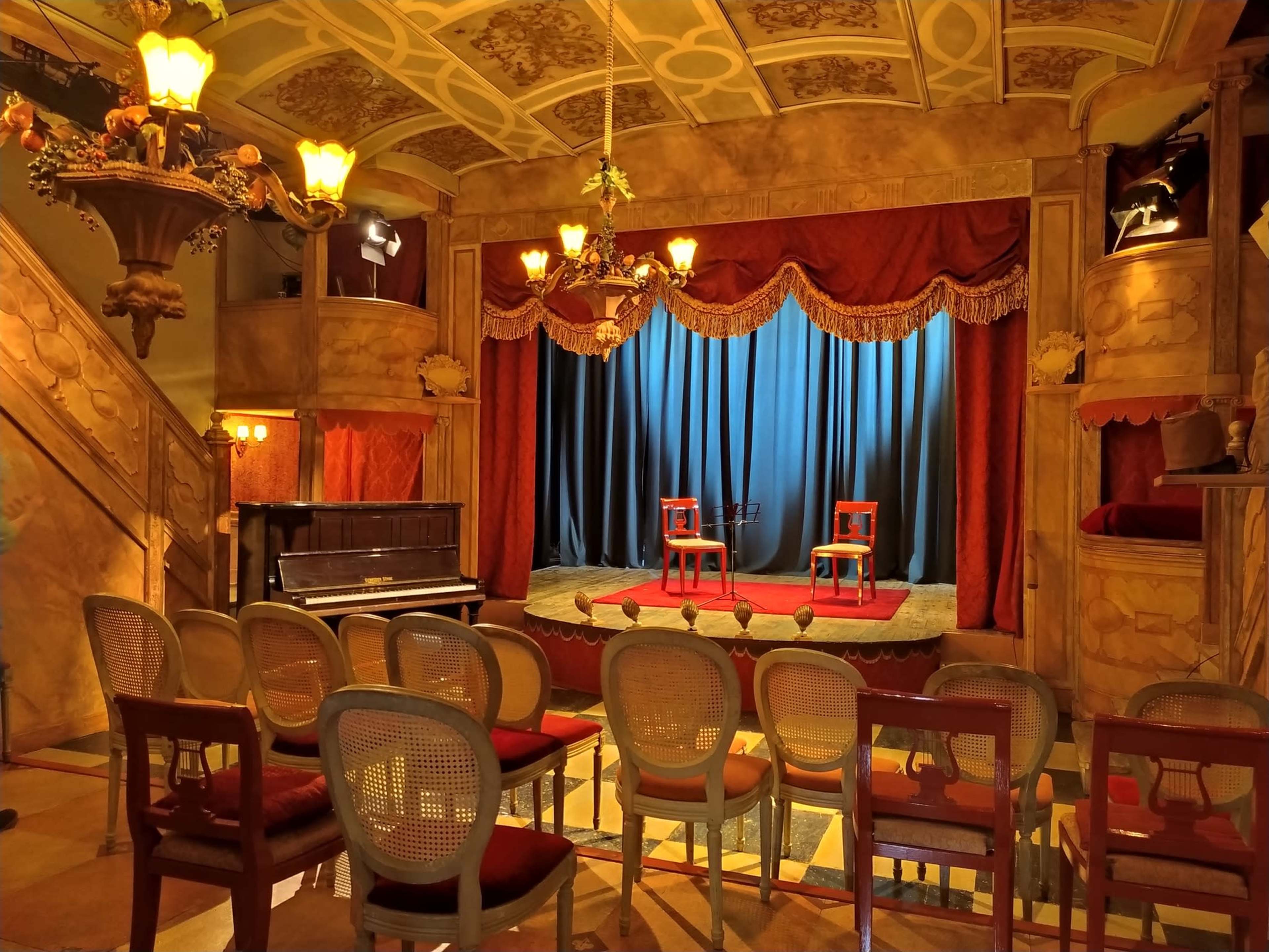 An empty theater stage with two chairs on a red carpet, flanked by a piano and a row of seats facing the stage.