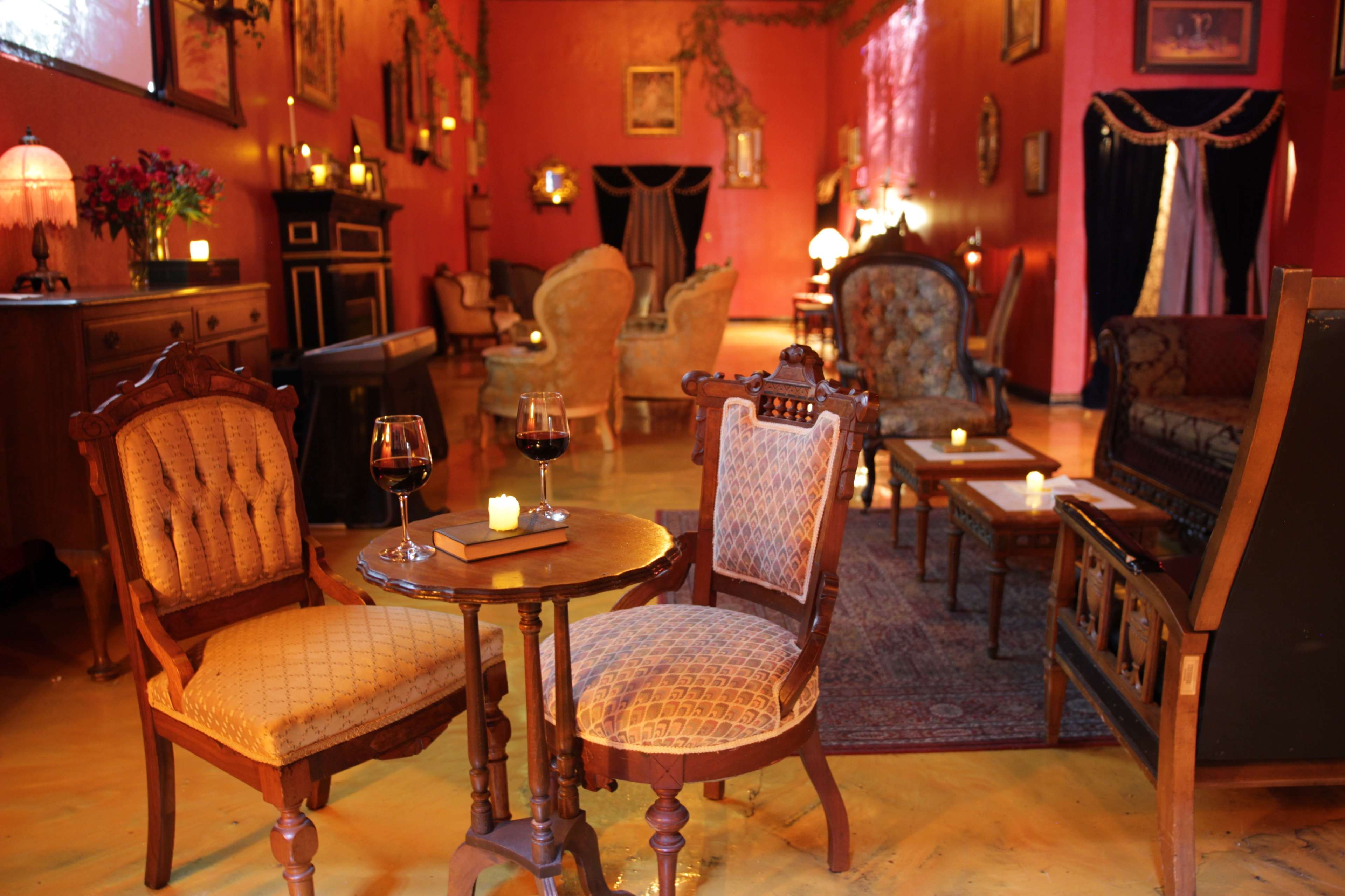 The scene features a cozy lounge area with vintage furniture, including two upholstered chairs, a small round table with glasses of red wine, and soft lighting from candles.