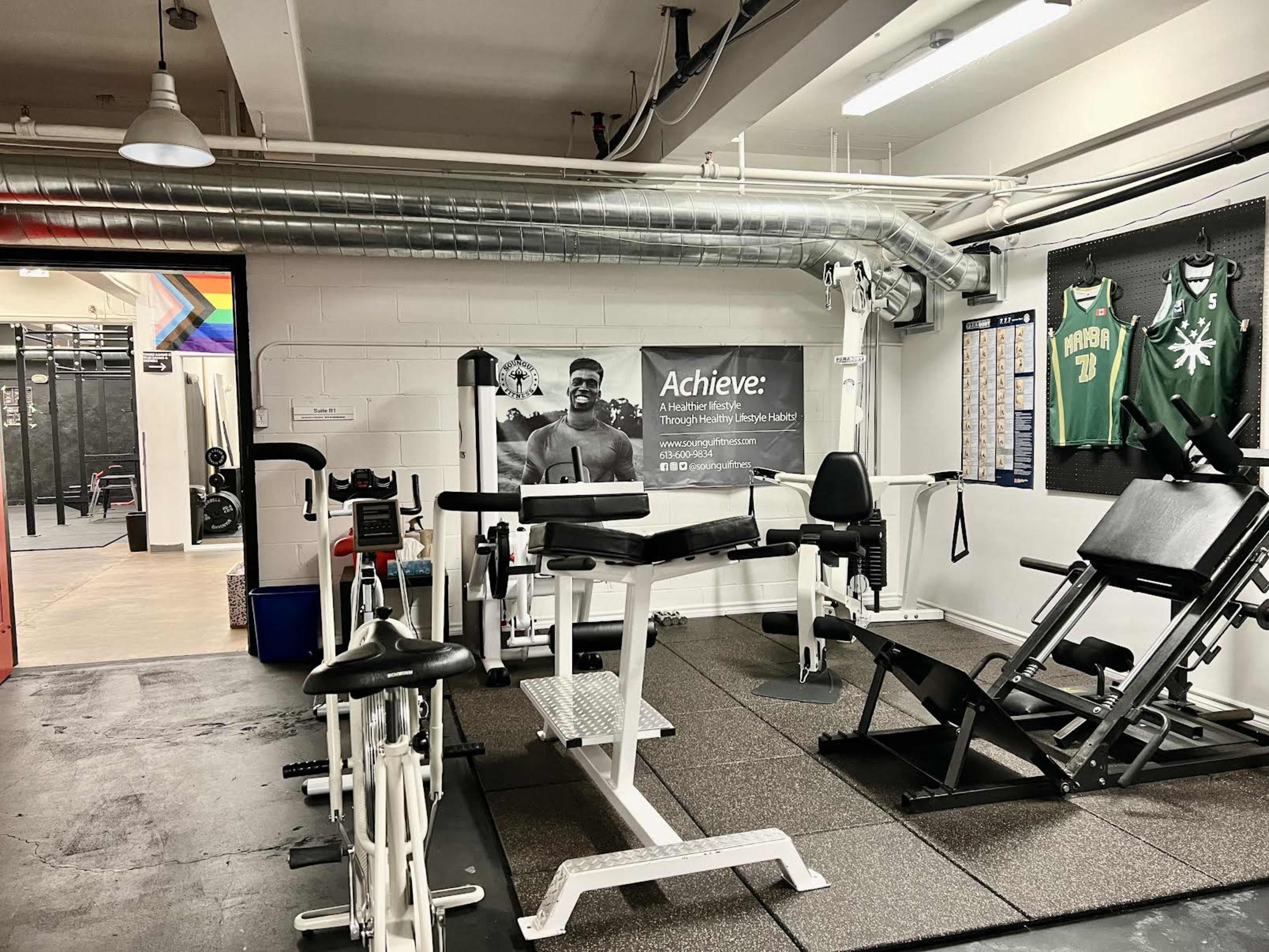 A gym area with various exercise machines and equipment, including a weight bench and resistance machines, adjacent to a wall featuring fitness posters and sports jerseys.
