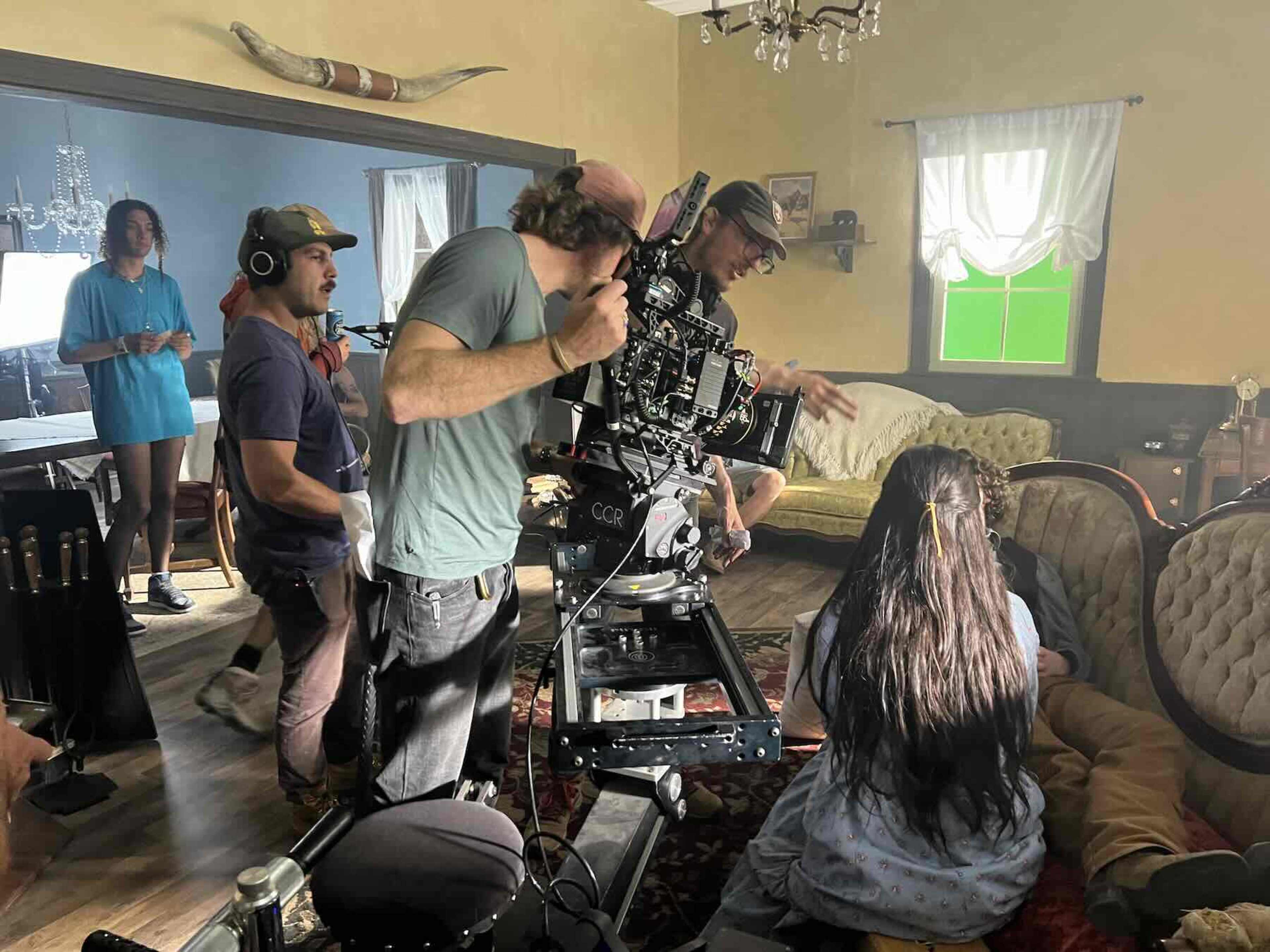A film crew operates a camera on a dolly inside a vintage-style living room while an actress sits on a couch.