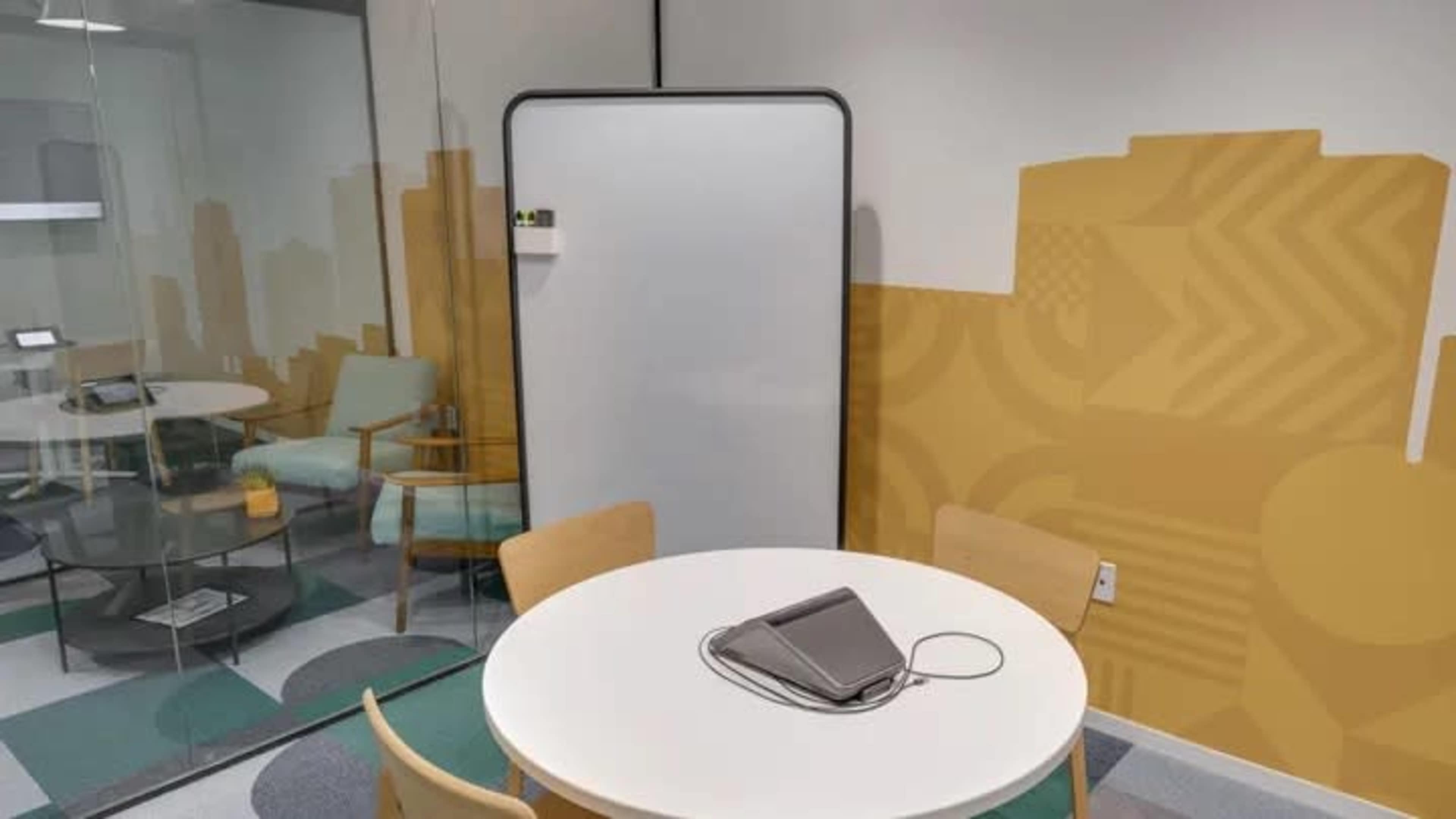 A small meeting room features a round table, chairs, a whiteboard, and colorful wall patterns.