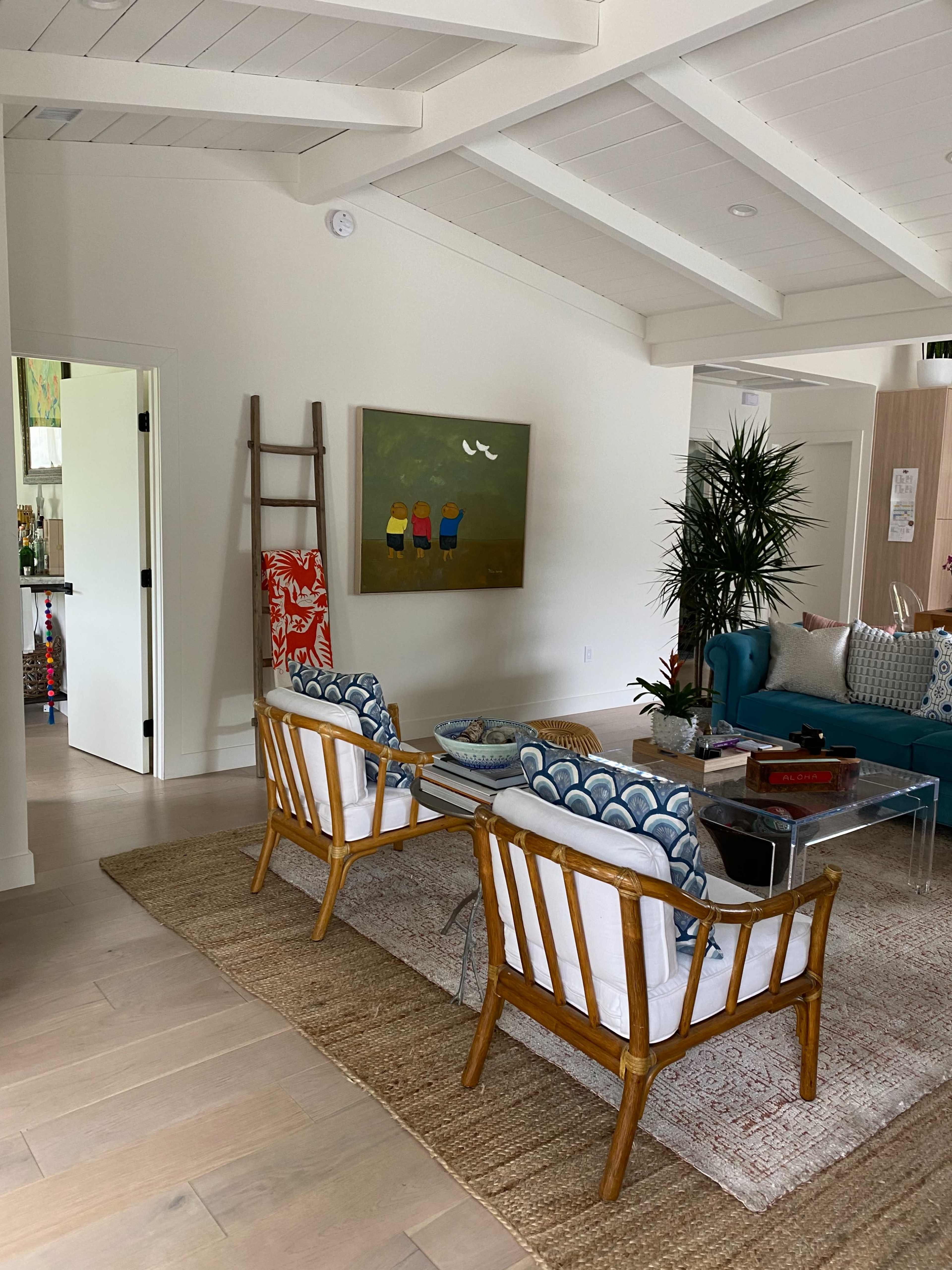 A brightly lit living room with wooden beams, featuring two rattan chairs with patterned cushions facing a glass coffee table, a green sofa, and a painting on the wall.