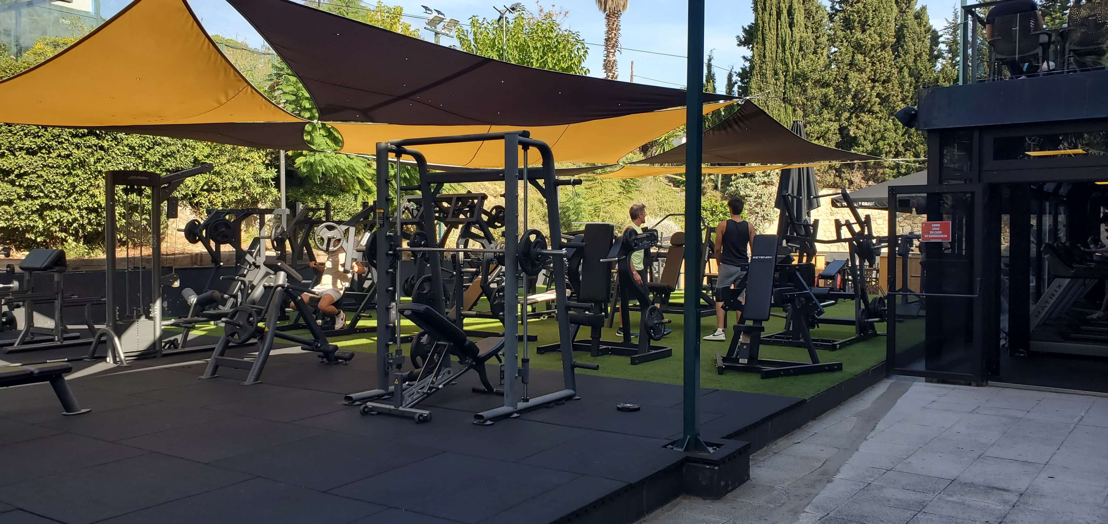 An outdoor gym area features various weightlifting and exercise machines under shaded canopies, with people actively working out.