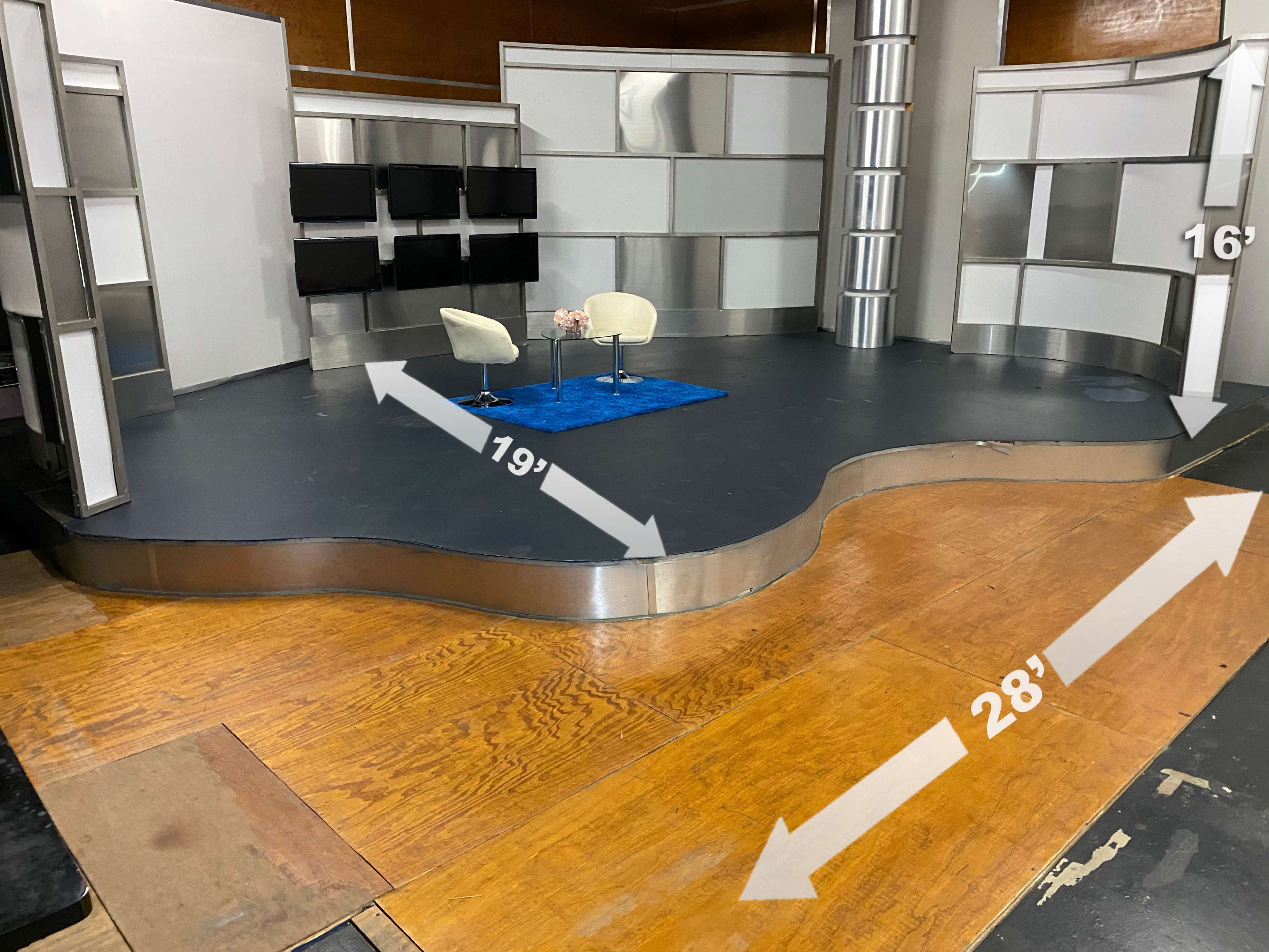 The image shows a television studio set featuring a curved stage with a blue carpet and two white chairs, surrounded by several flat-panel screens.